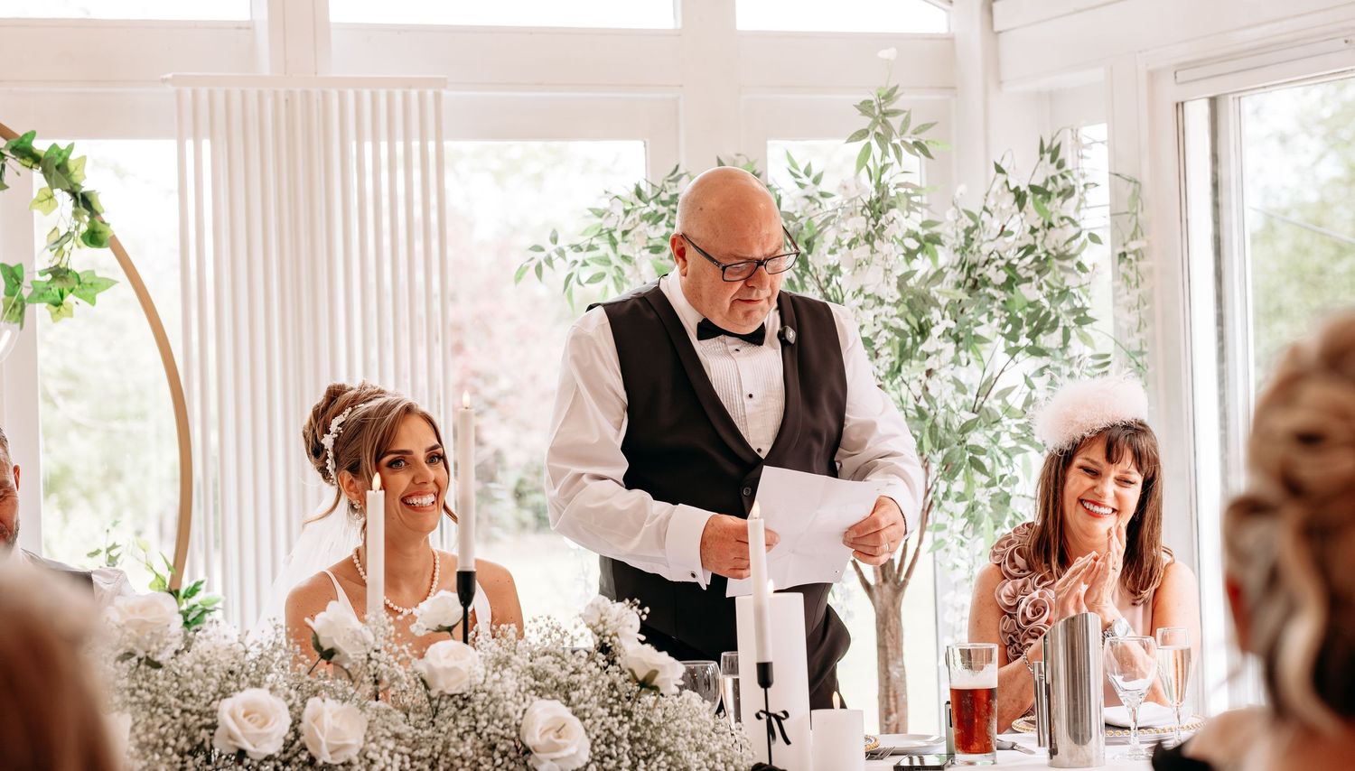 Elderly man in vest giving wedding speech at head table with bride and guests laughing in elegant venue.