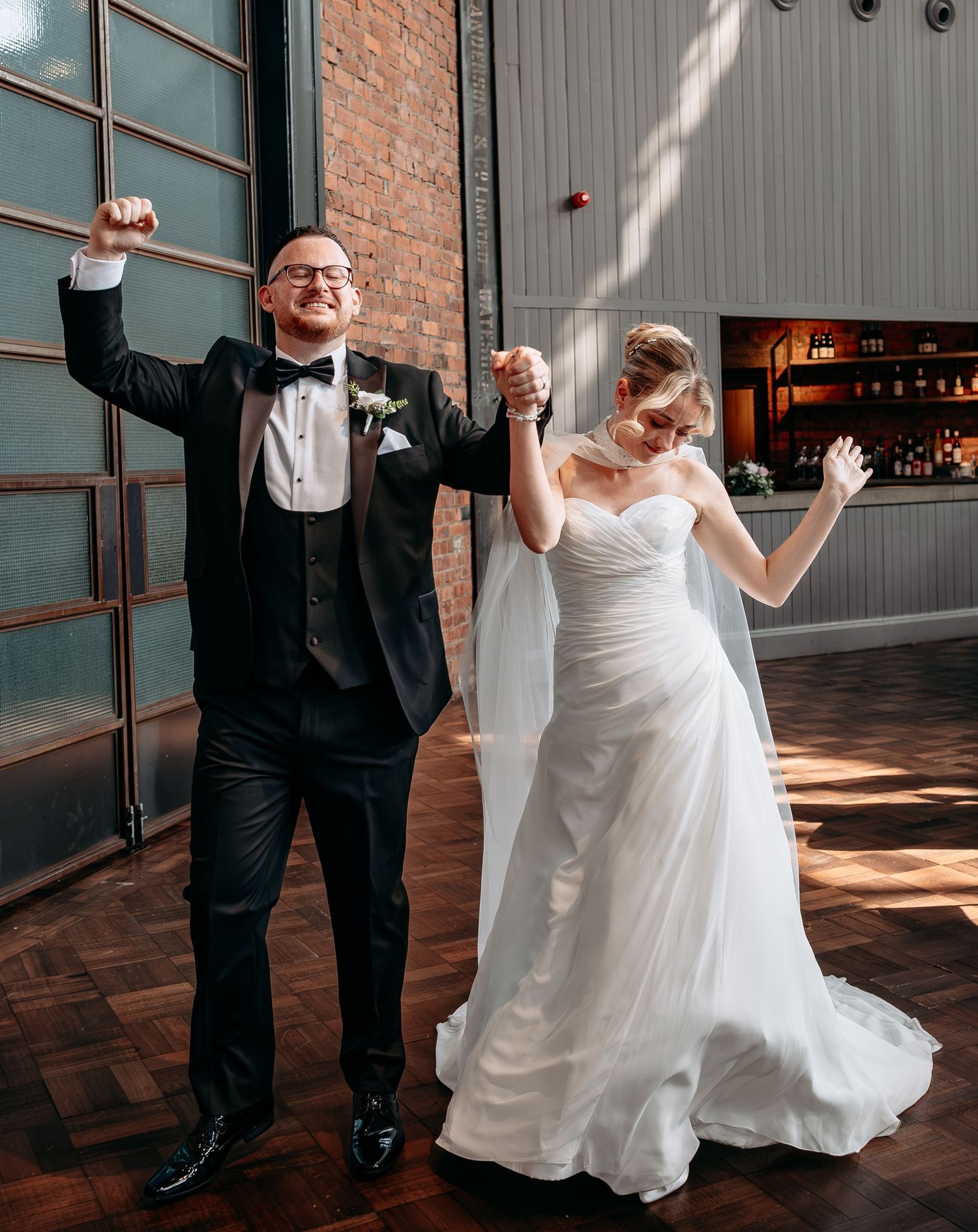 Joyful newlyweds celebrate their grand entrance, groom in tuxedo raising fist, bride in white gown holding his hand.
