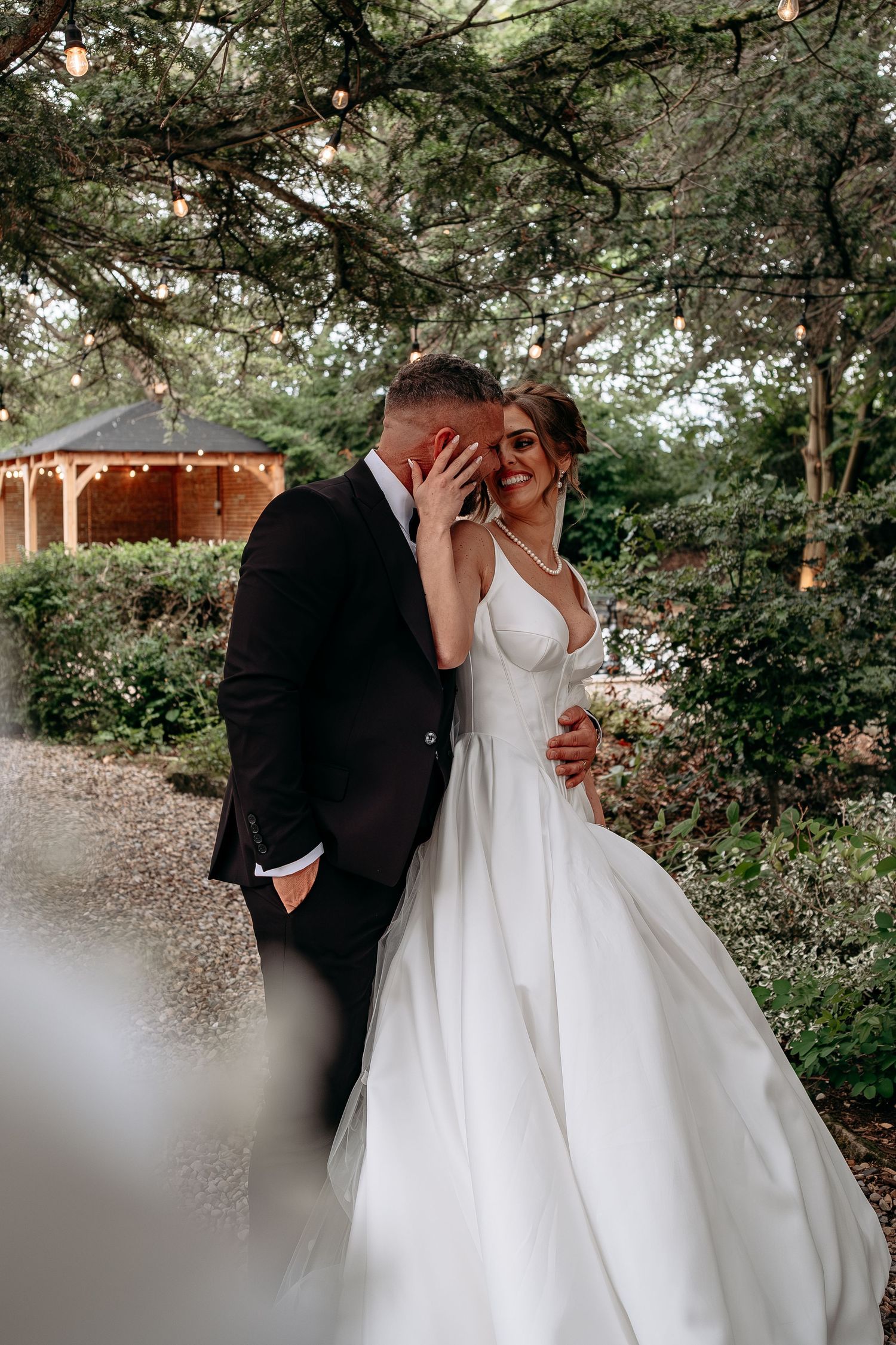 Bride in white gown and groom in black suit share a romantic moment in a lush garden setting.