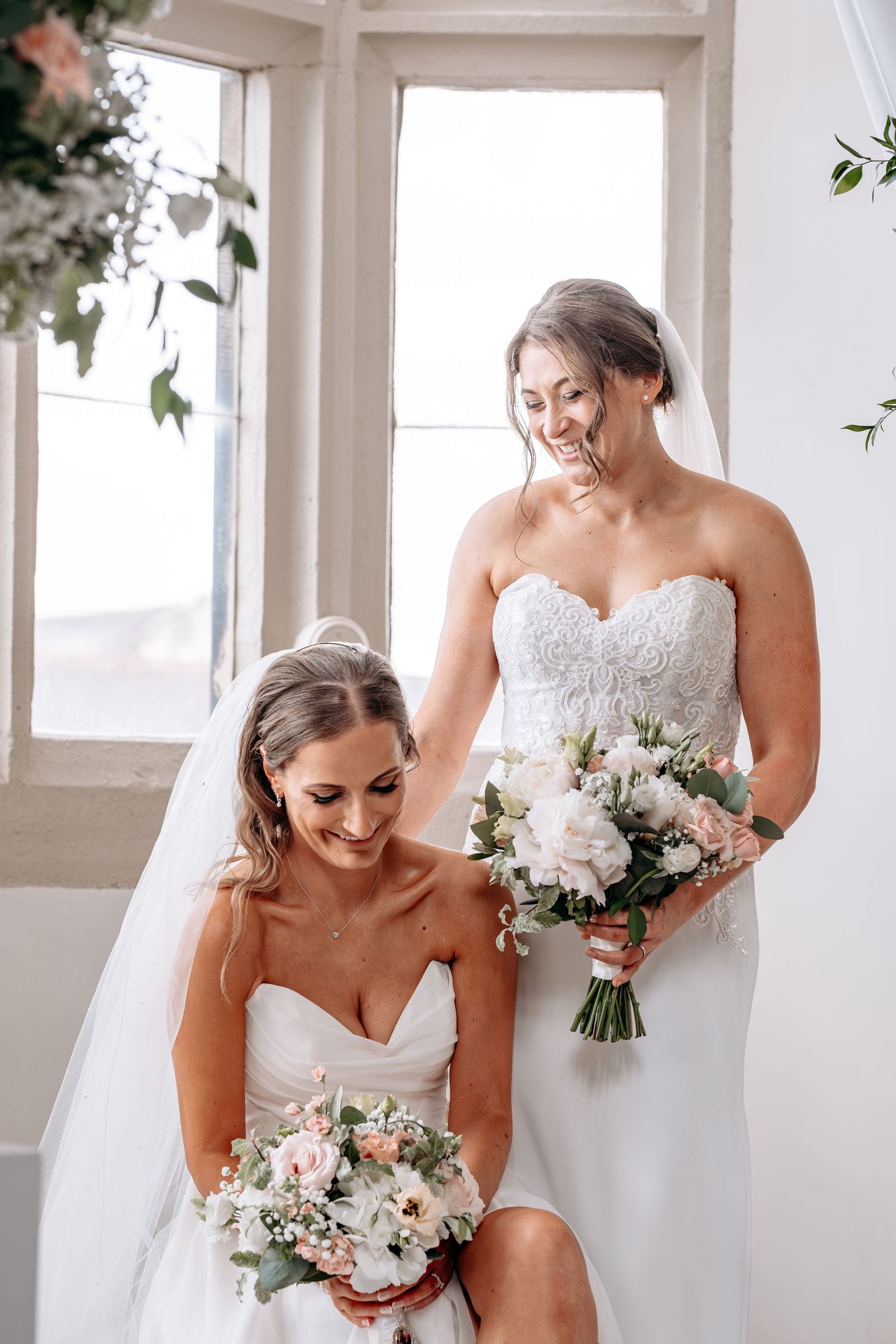 Two brides in strapless white gowns holding bouquets of blush and white flowers, smiling together near a bright window.