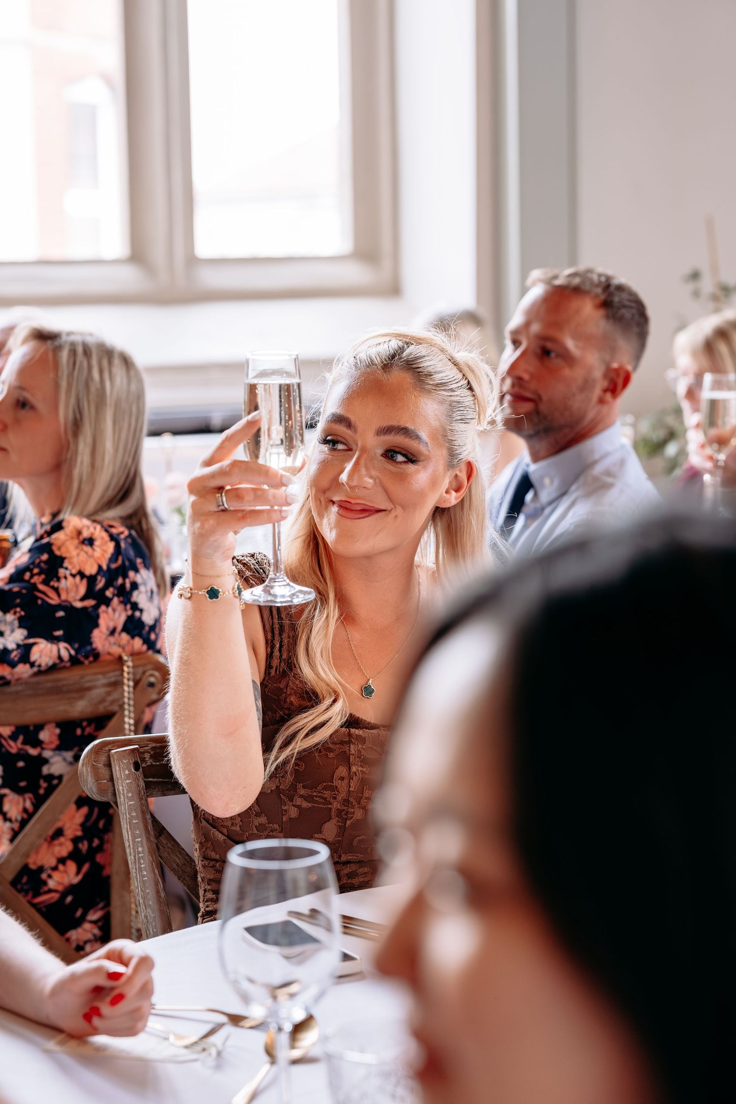 Blonde woman raising a champagne glass toast at an elegant wedding reception dinner, surrounded by guests at a decorated table.