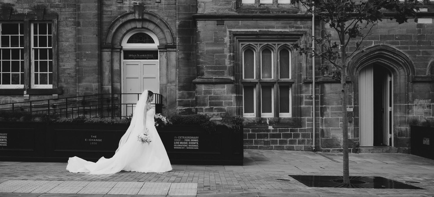 Black and white photo of a bride in a flowing white gown and veil walking outside a historic stone building.
