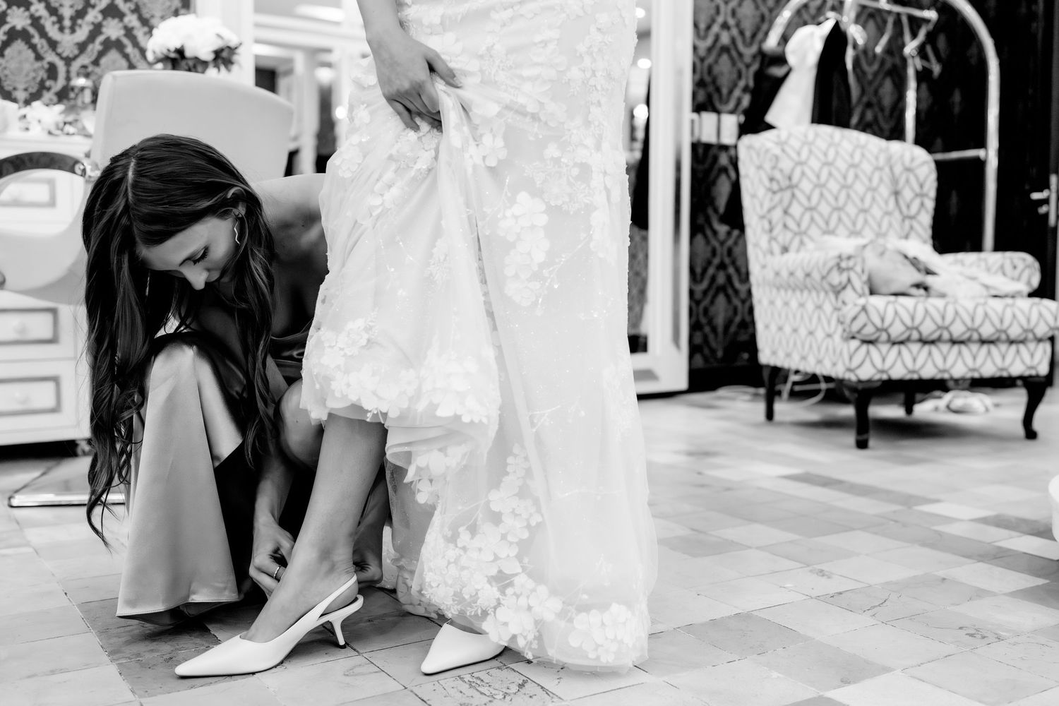 Bridesmaid kneels to help a bride slip into white pointed heels while holding her lace wedding gown in a bridal boutique.