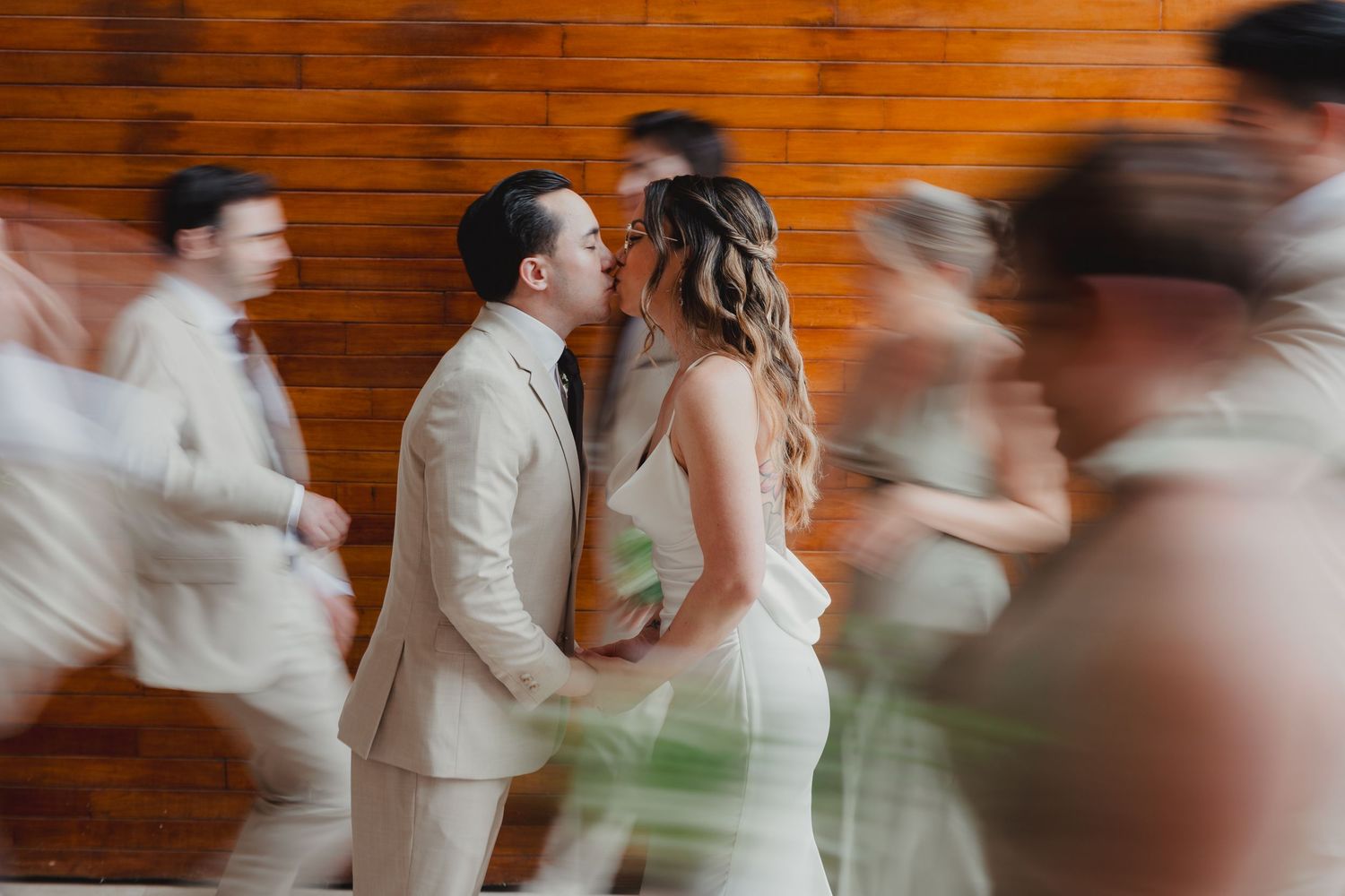 A bride and groom share a kiss while the wedding party moves around them in a motion-blurred photo.