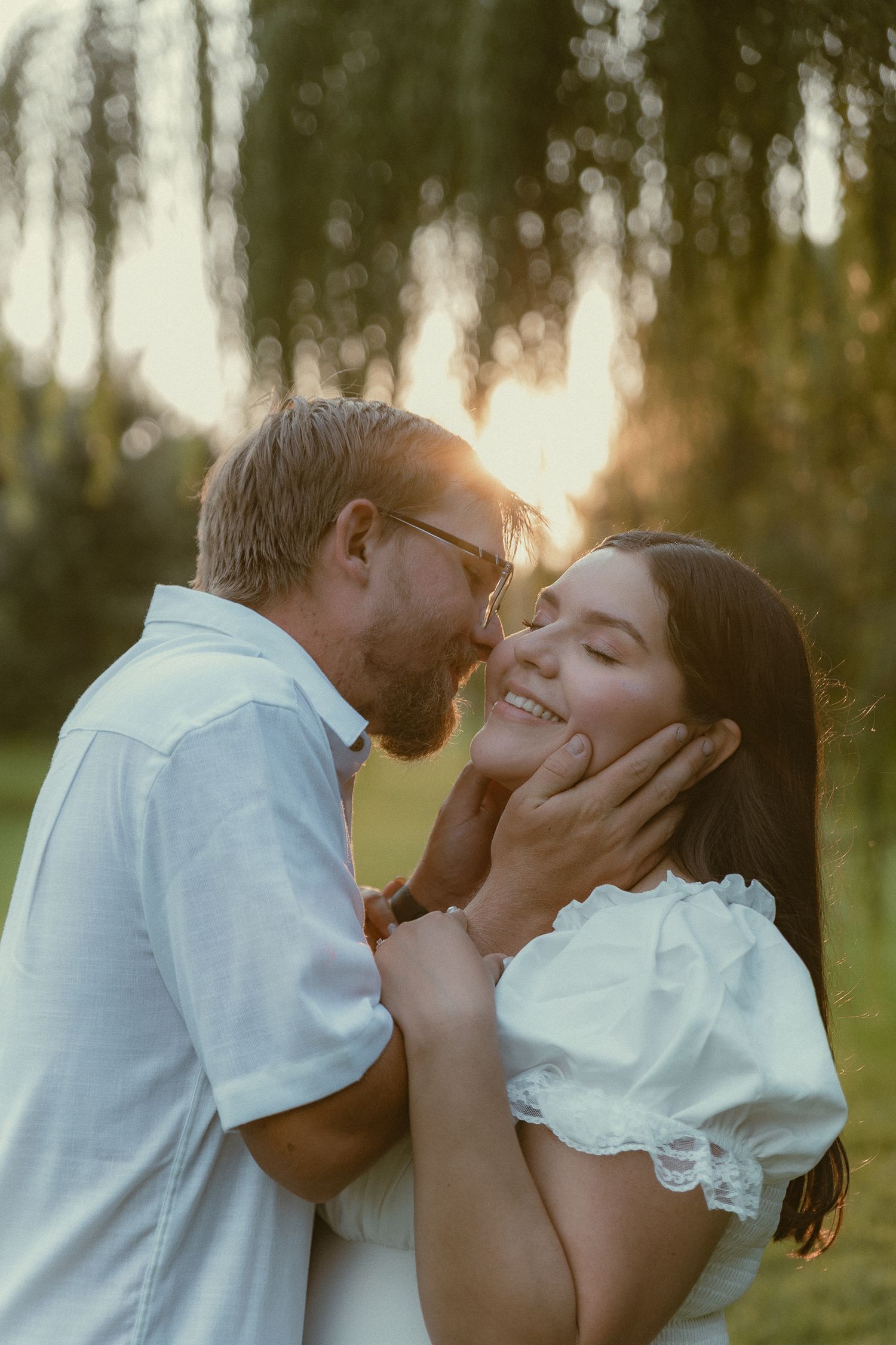 Couple sharing a romantic moment at golden hour, man tenderly holding woman's face as she smiles with eyes closed outdoors.