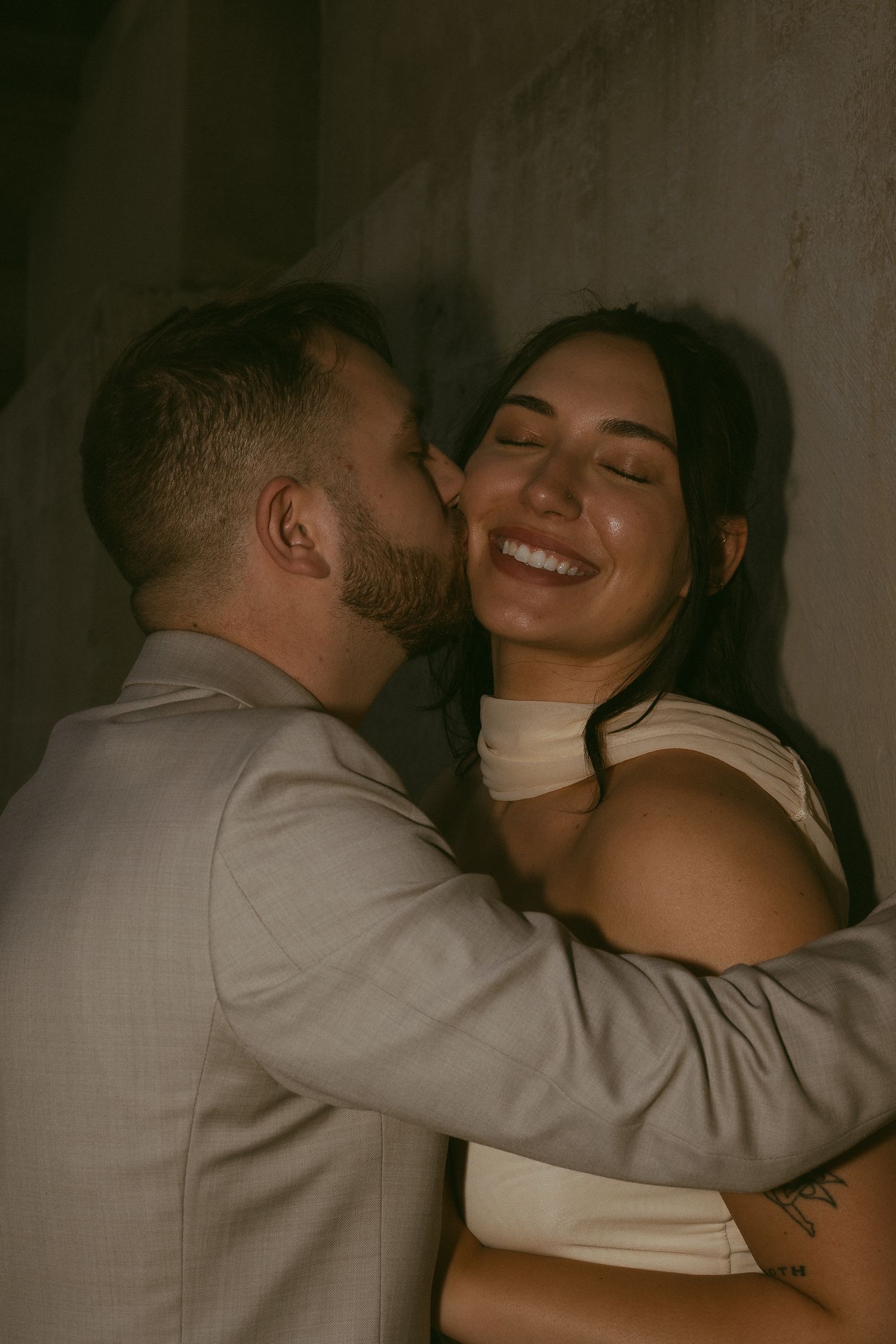 A couple shares a romantic embrace, man kissing woman's cheek as she smiles joyfully against a dark wall.