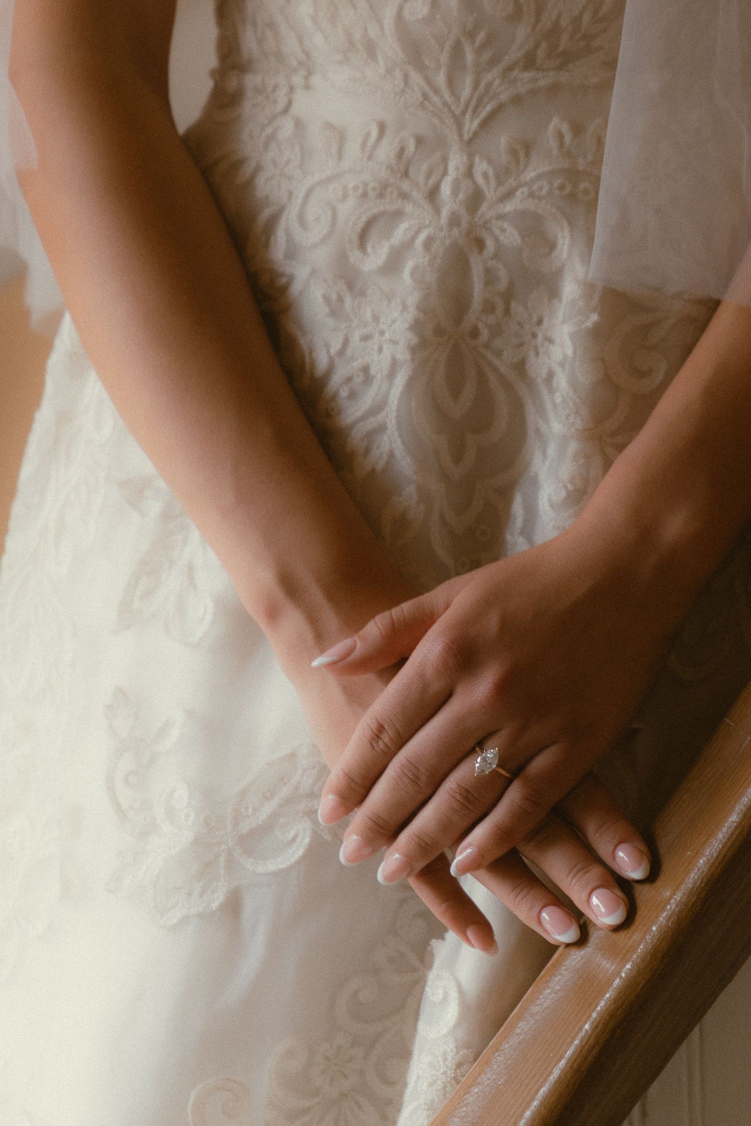 Bride in lace wedding dress with elegant French manicure and diamond engagement ring resting hands on wooden railing.