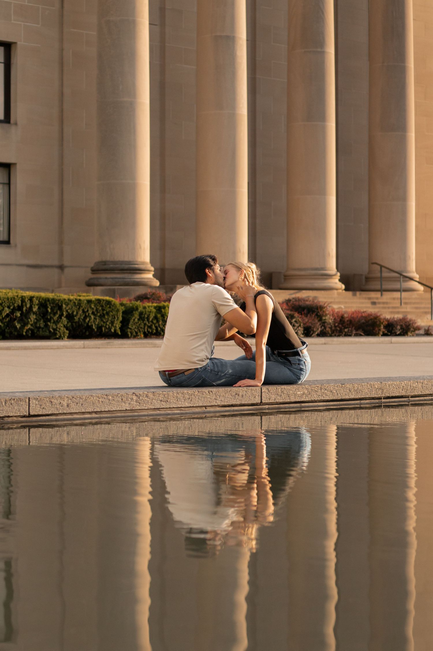 Couple sharing a romantic kiss beside a reflective pool in front of a classical building with tall stone columns.