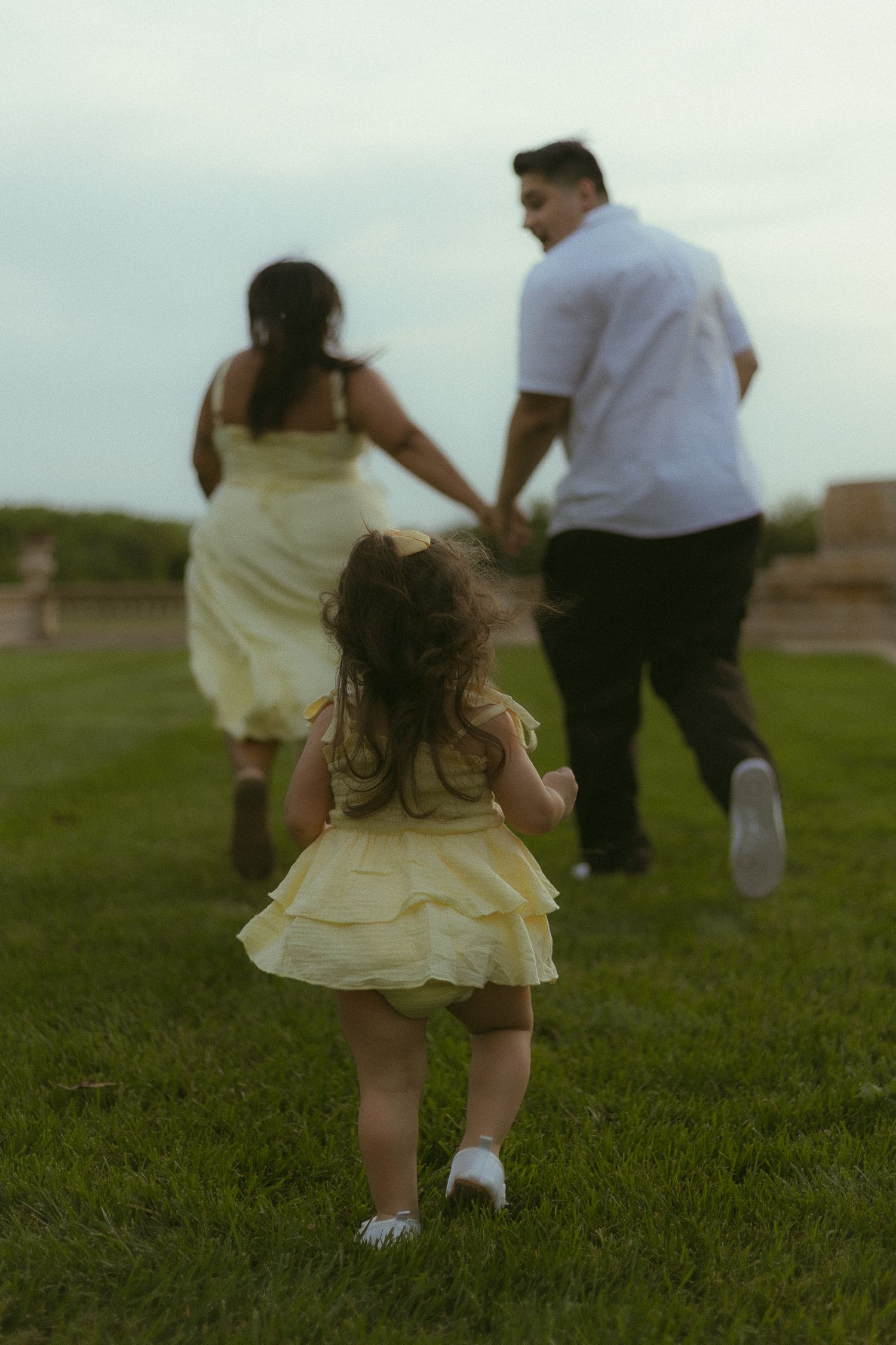 A little girl in a yellow dress runs toward her parents holding hands on a green grass field.