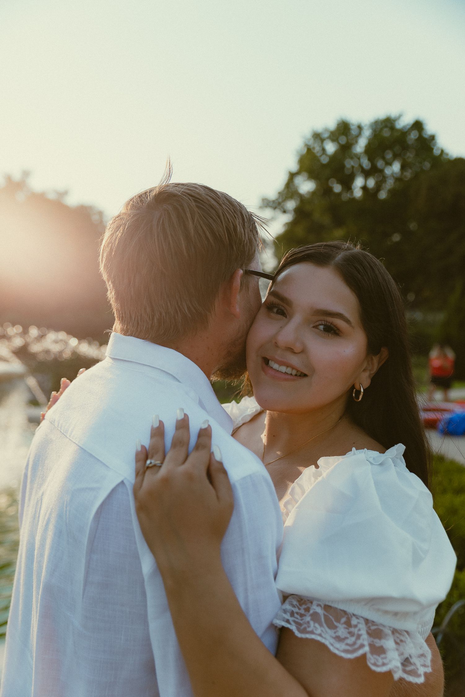Couple embracing at golden hour, woman in white lace top smiling, man kissing her cheek outdoors.