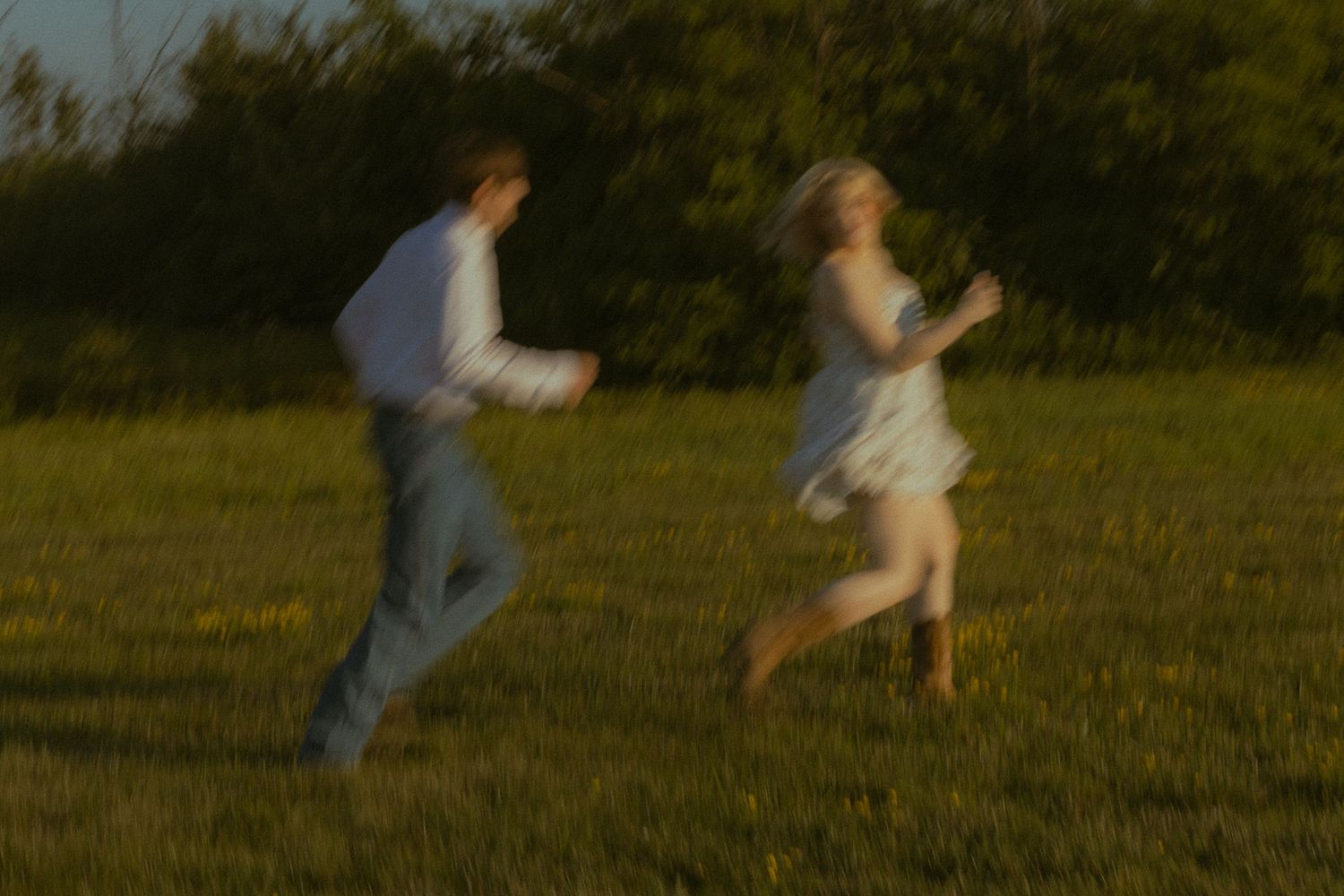 A blurry motion shot of a young couple running together through a sunlit green field at dusk.