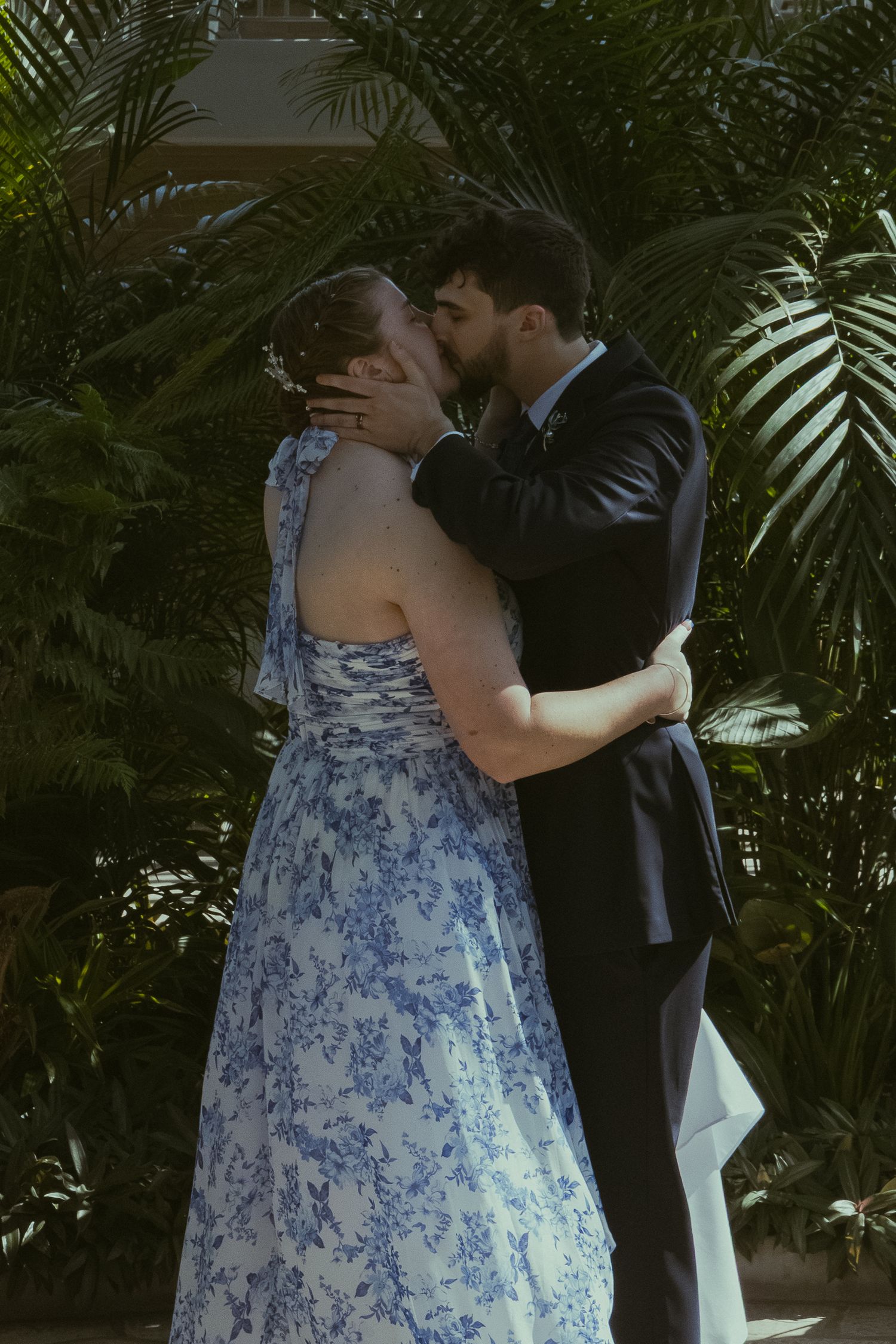 A couple shares a romantic kiss, the woman in a blue floral dress and the man in a dark suit, surrounded by lush greenery.