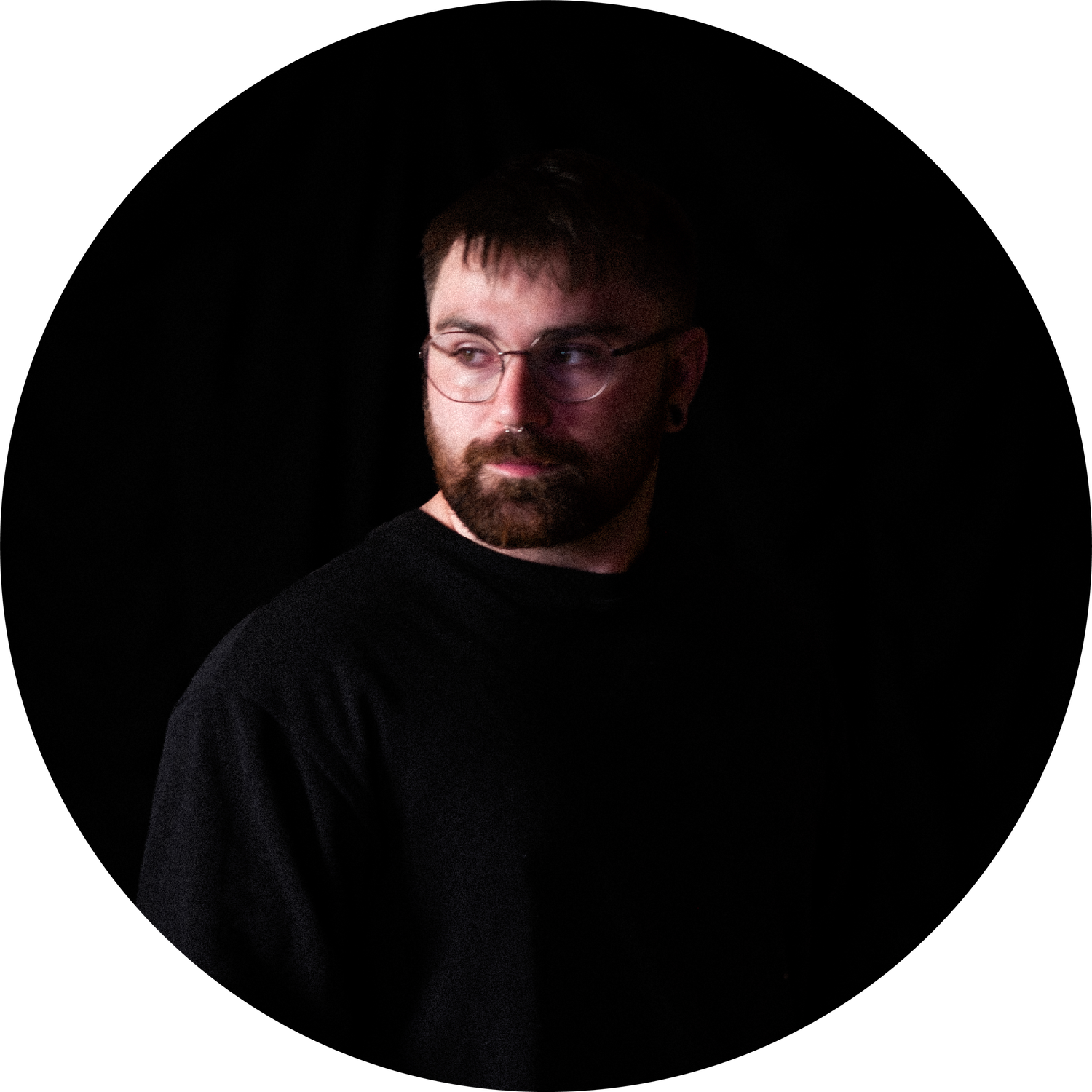 Bearded man with glasses wearing a black sweater, photographed in dramatic low-key lighting against dark background.