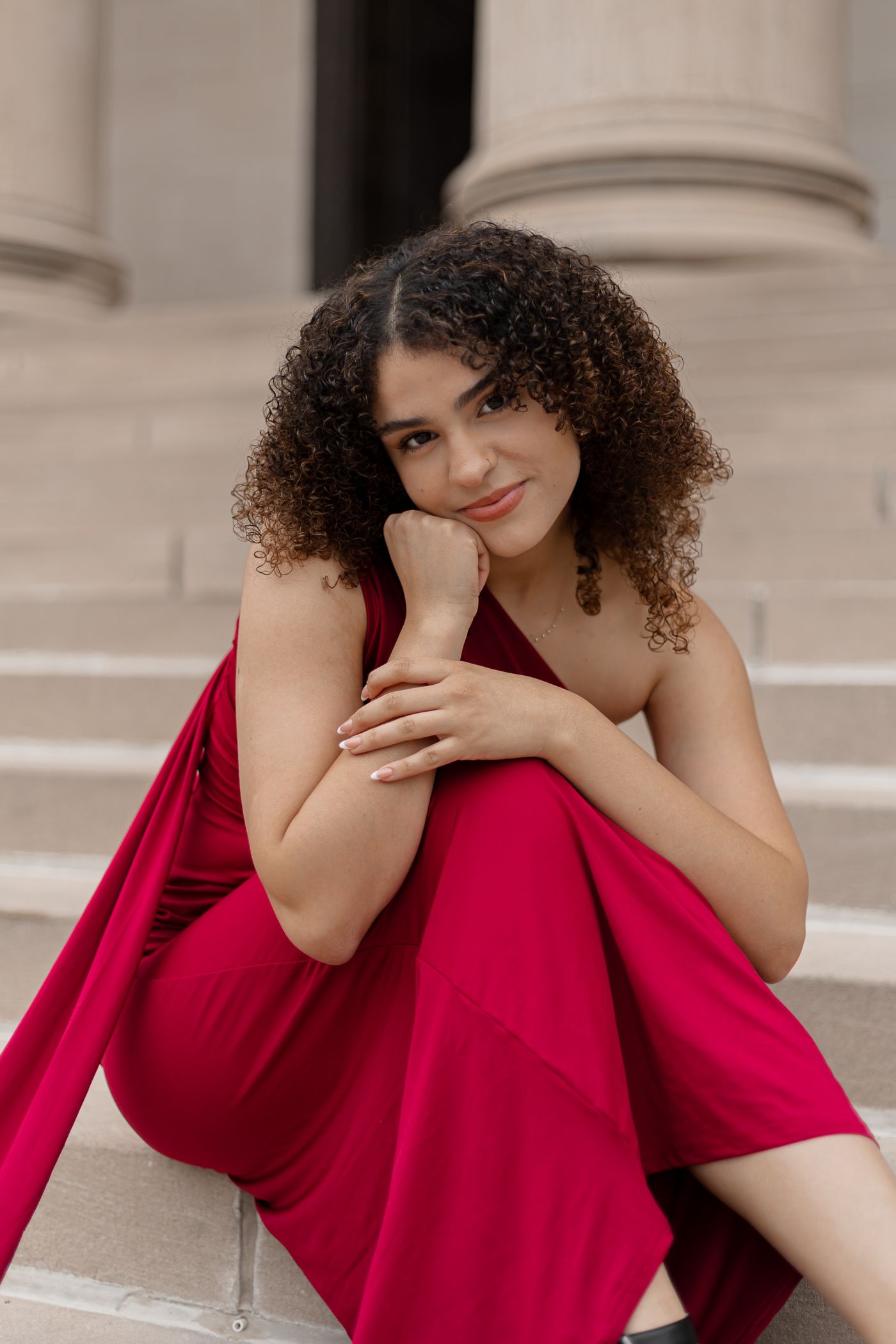 A woman in a vibrant red dress poses elegantly against a classical architectural backdrop with columns.