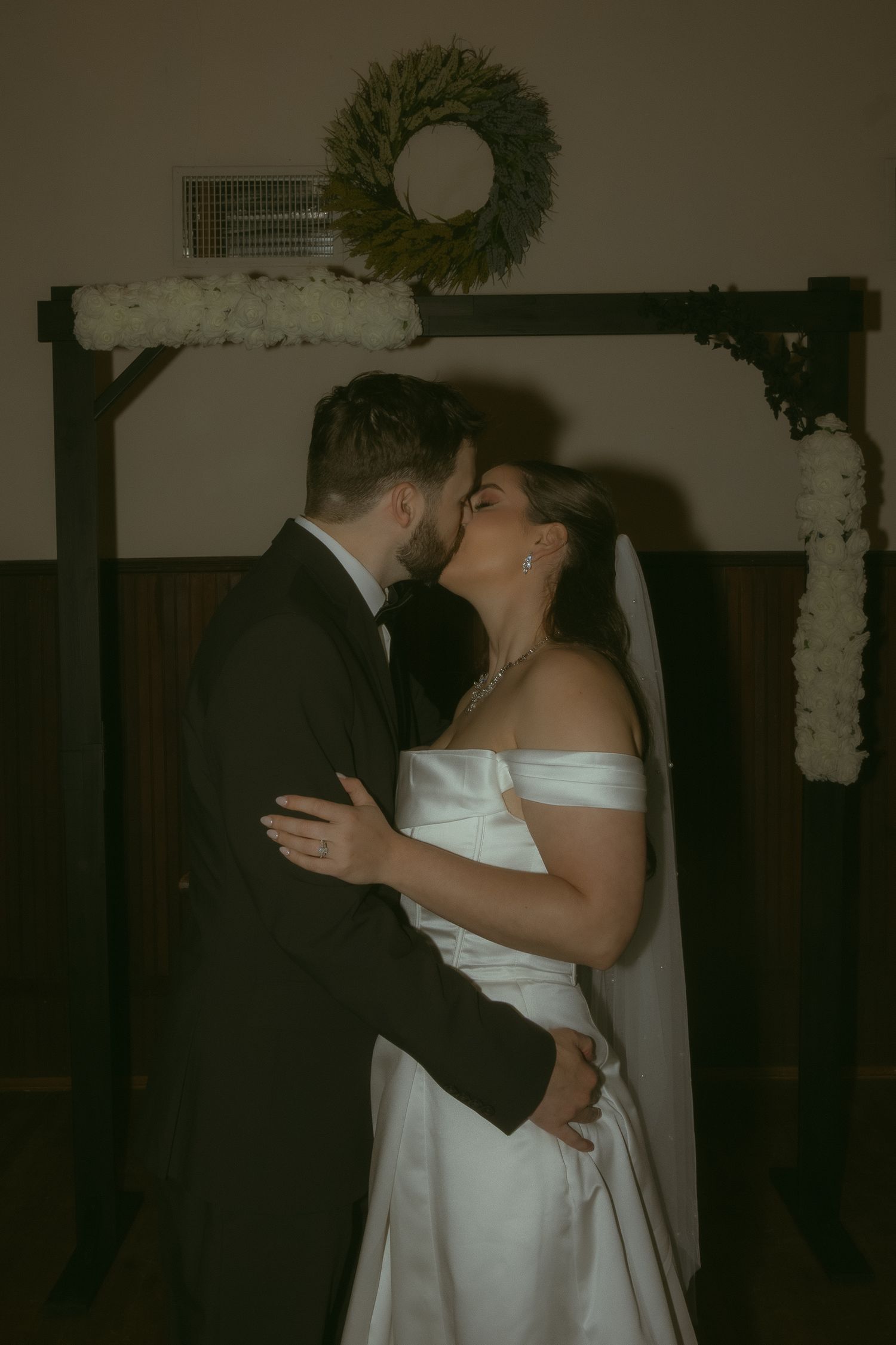 Bride and groom share their first kiss under a floral arch adorned with white flowers and a green wreath.