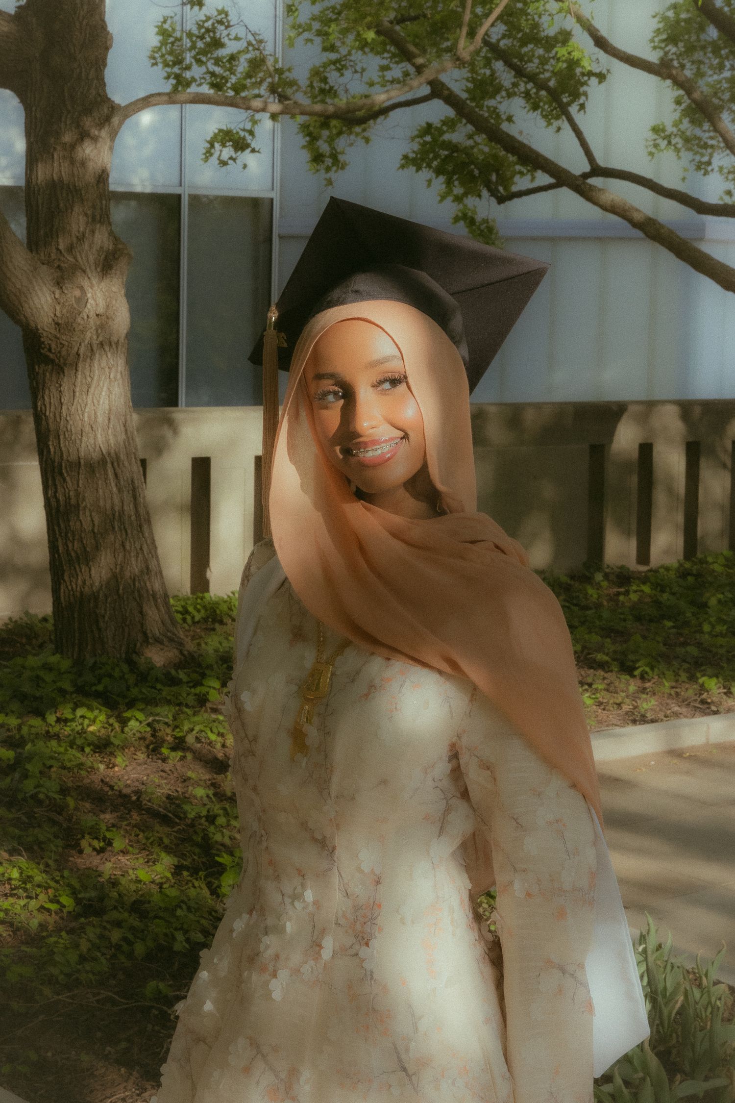 Smiling graduate in floral dress and black cap with blonde hair and hijab posing outdoors near trees.