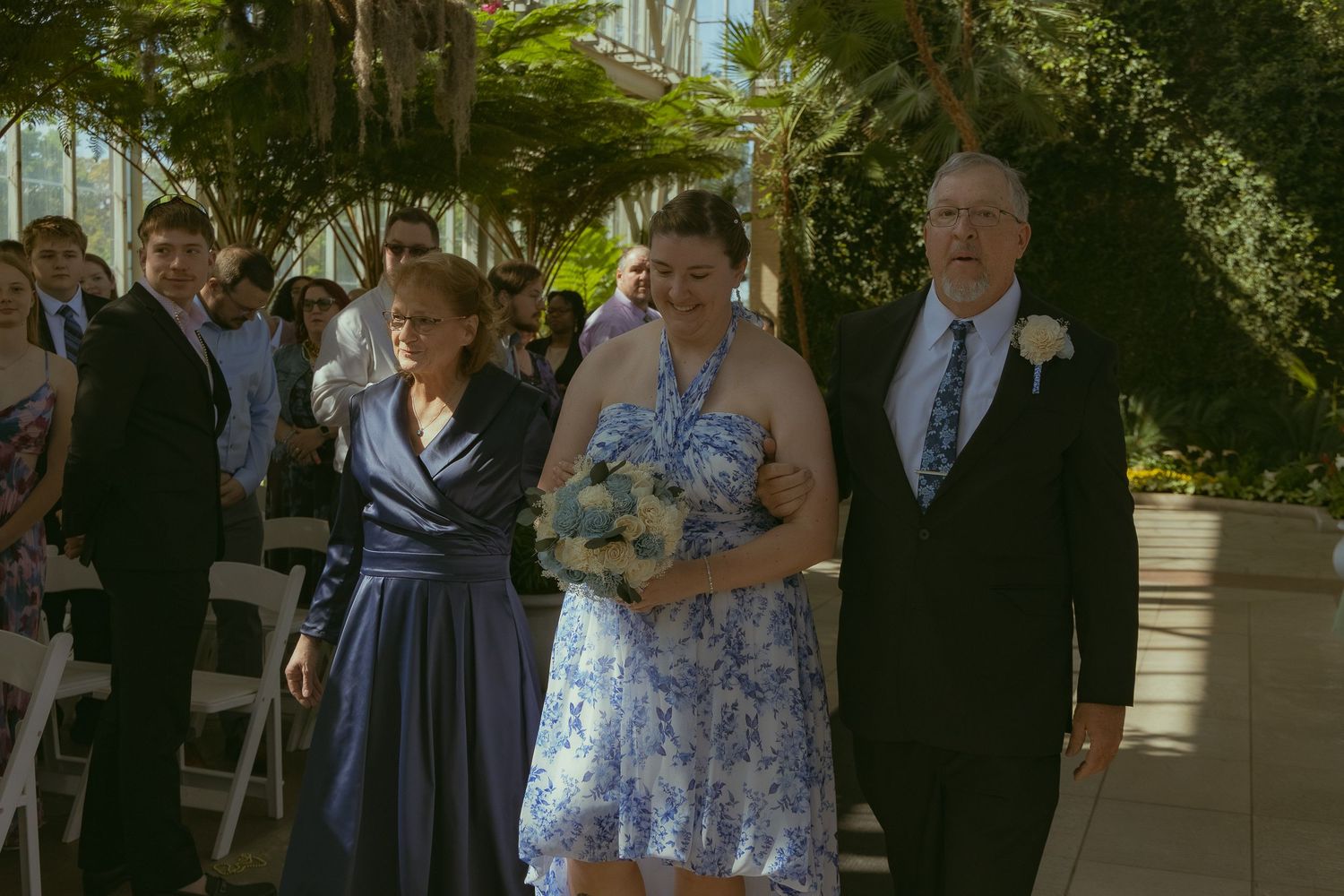 Bride in blue floral dress walks down outdoor wedding aisle with parents, guests seated among lush greenery.