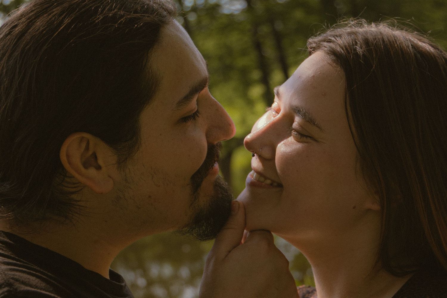 Romantic couple sharing an intimate moment outdoors, smiling and touching noses in a sunlit forest setting.