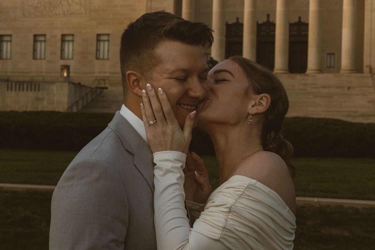 A couple shares a romantic kiss outdoors, the bride in white and groom in gray suit, with a grand building backdrop.