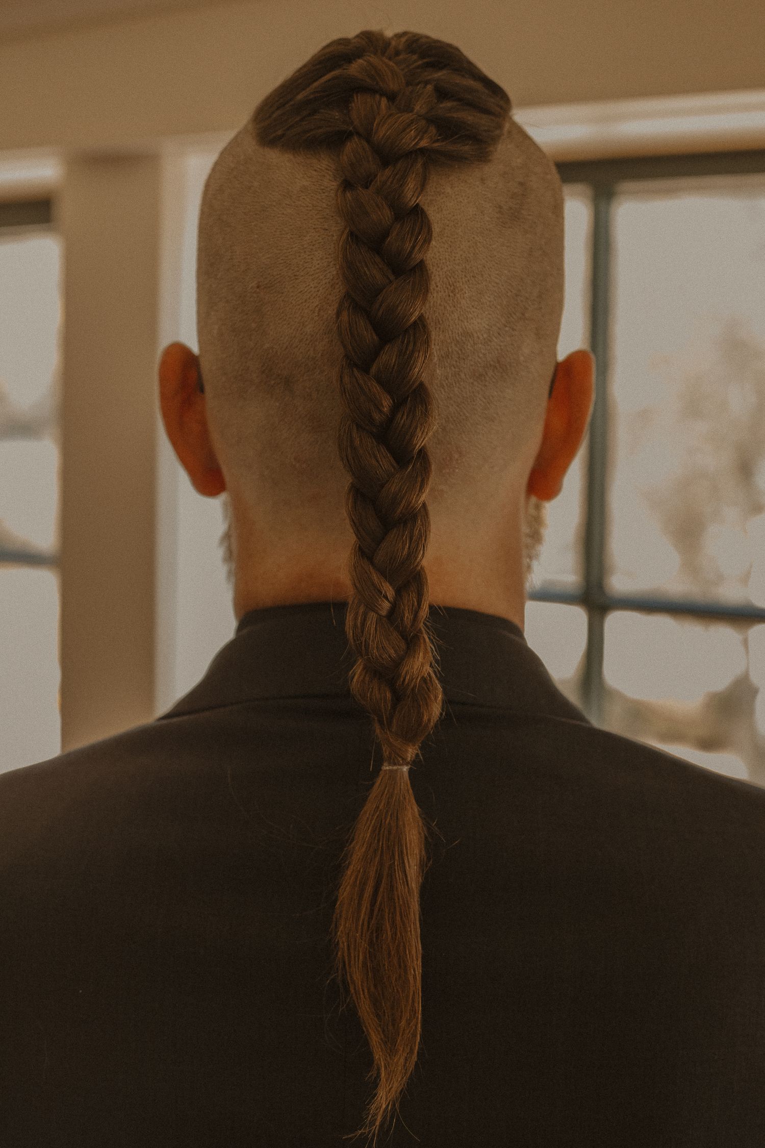 Back view of person with long braided hair wearing dark clothing against a light background.