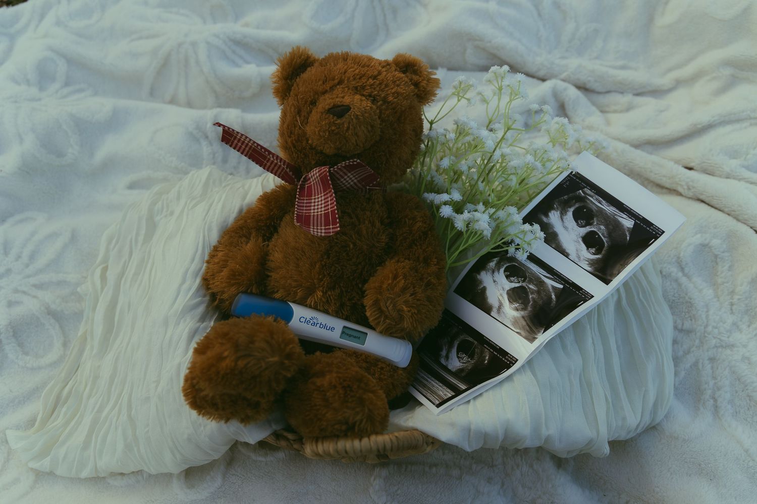 Brown teddy bear with plaid bow beside pregnancy test, ultrasound photos, and white flowers on white lace fabric.