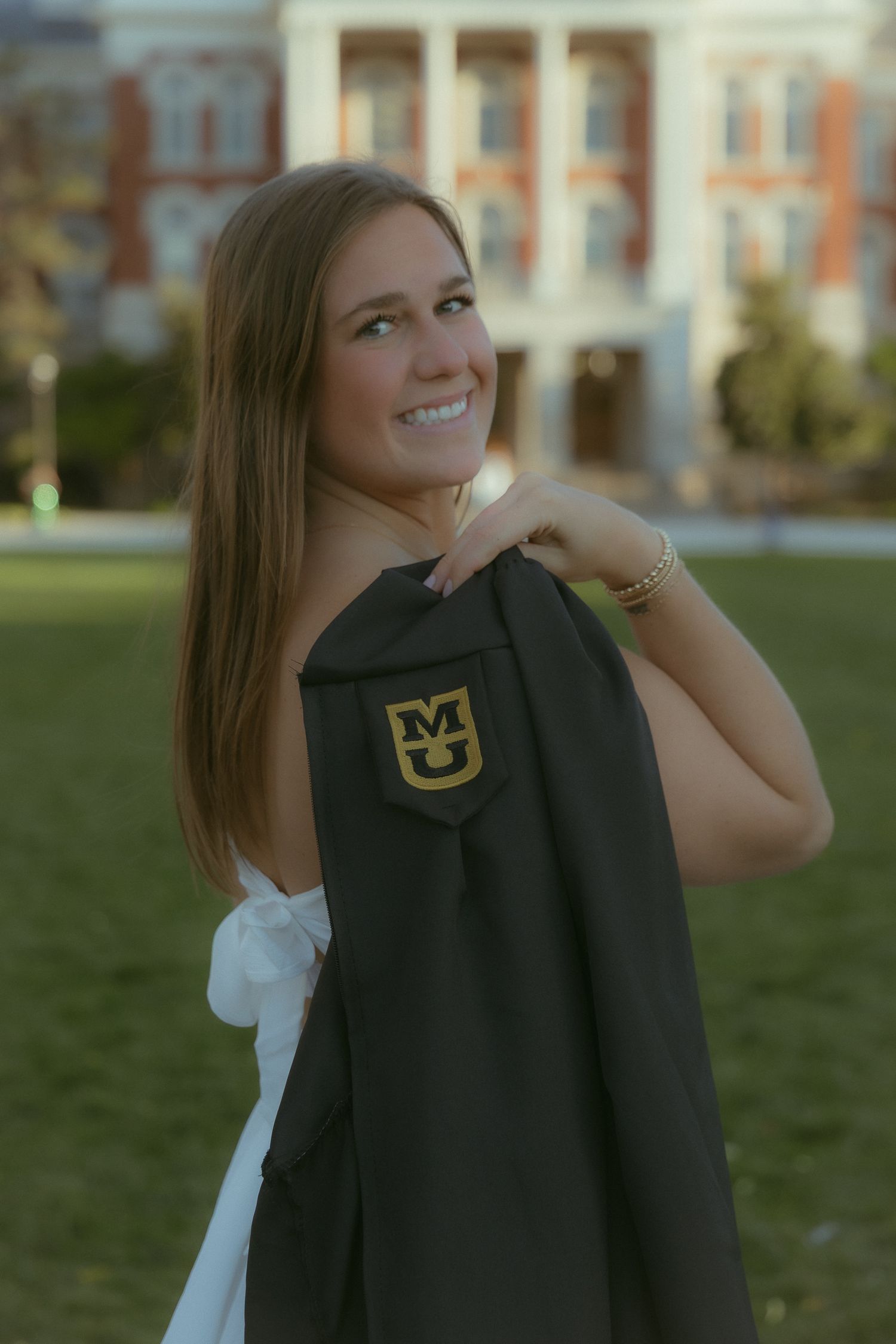 Smiling young woman holding a University of Missouri graduation gown over her shoulder outdoors on campus.