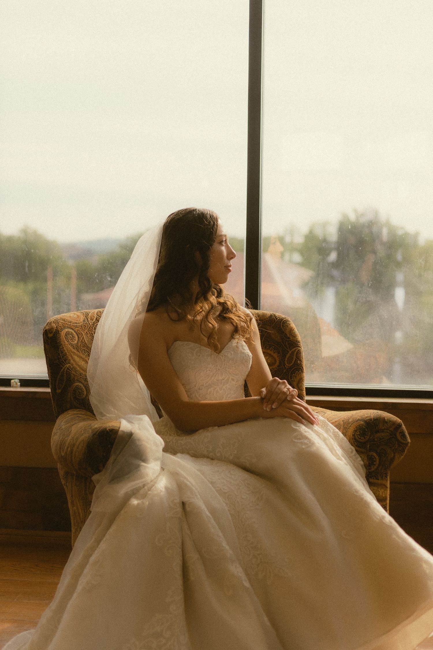 Bride in white lace gown and veil sits in ornate chair gazing out large window on wedding day.