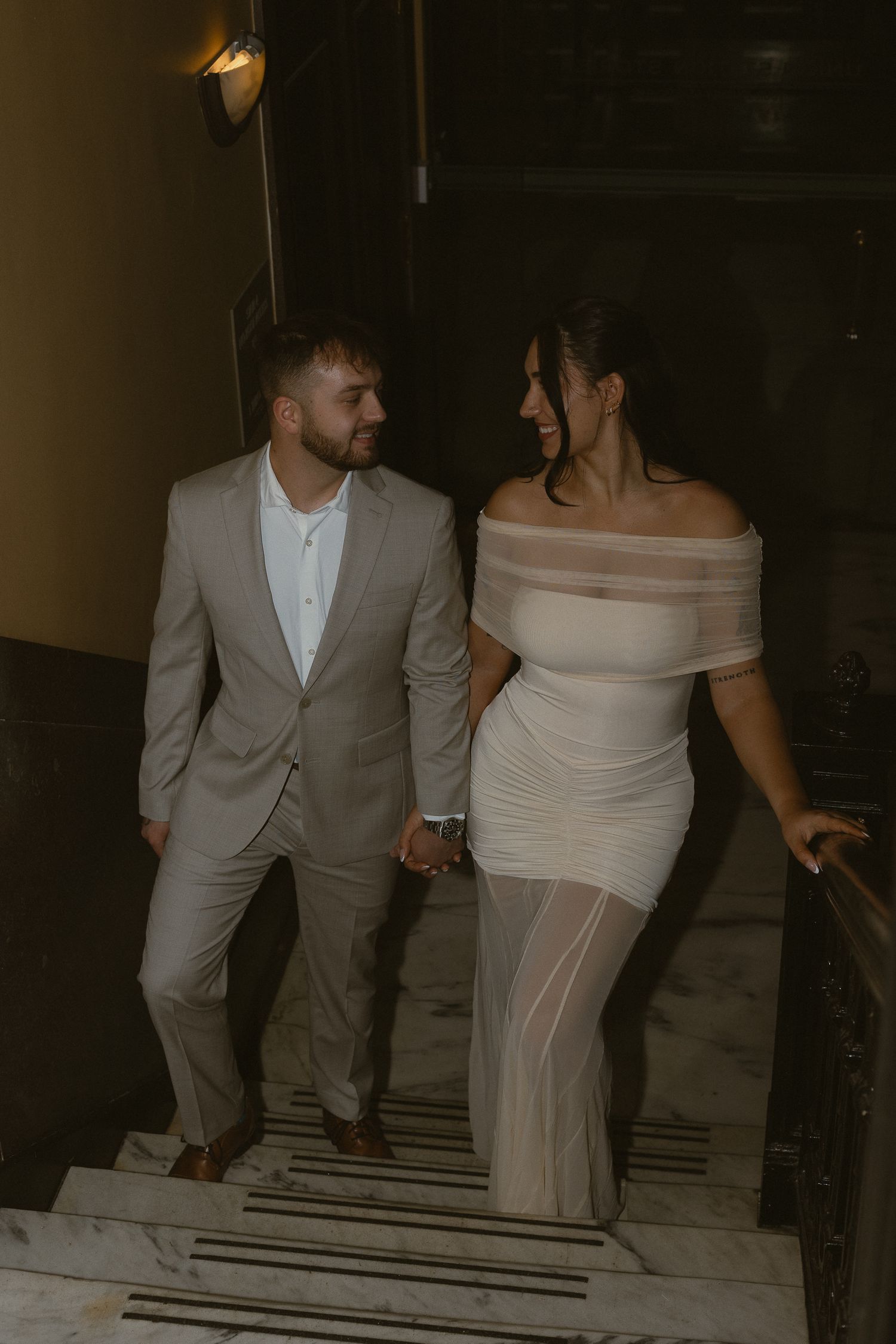 Couple in formal attire holding hands on marble stairs, man in gray suit, woman in white off-shoulder dress.