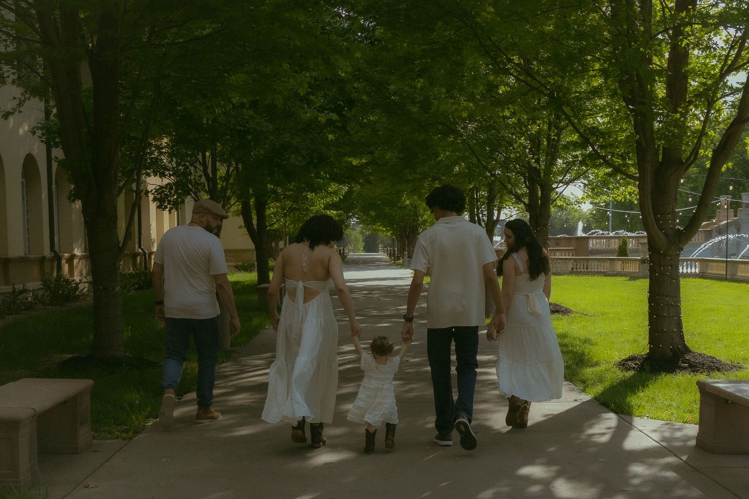 Family of five walking hand-in-hand down a tree-lined path, dressed in white outfits on a sunny day.