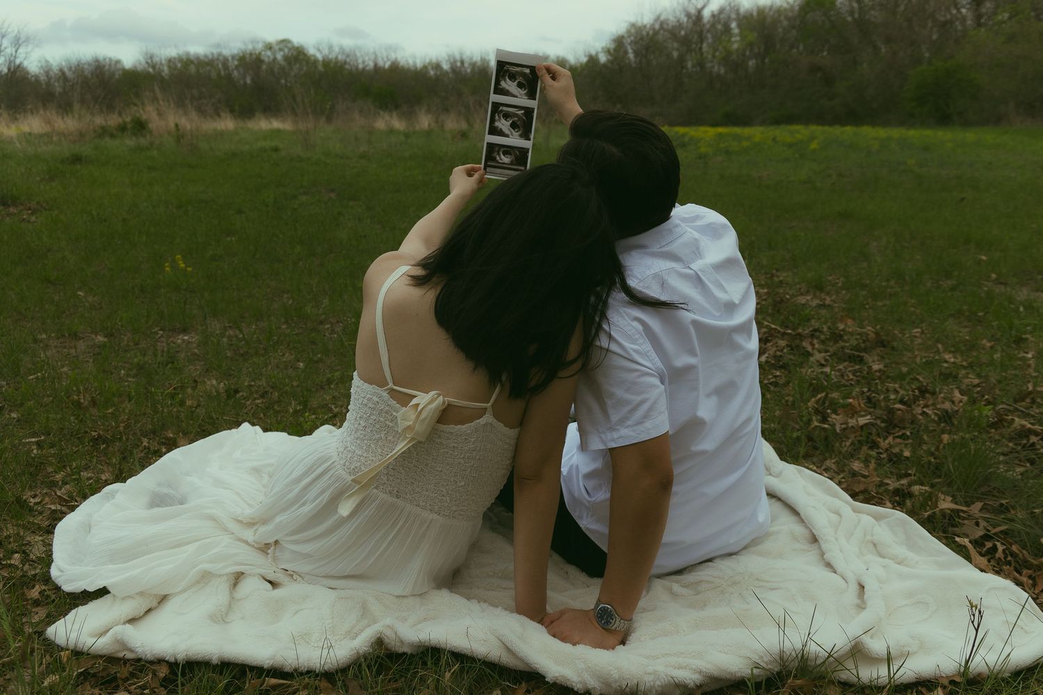 Couple sitting on blanket in field holding ultrasound photo, pregnancy announcement maternity outdoor portrait.
