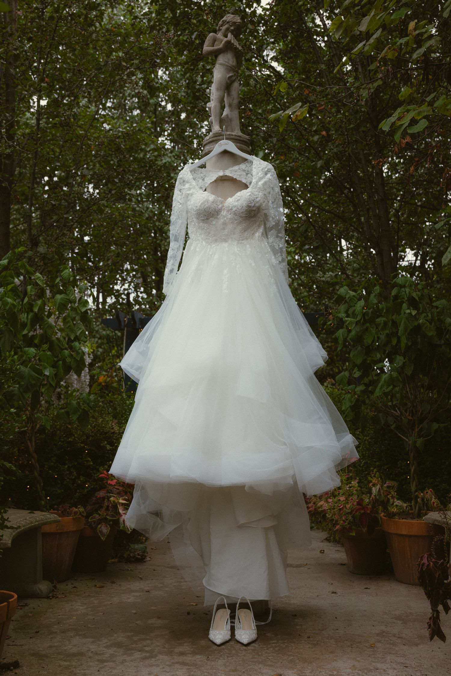 White lace long-sleeve wedding dress with layered tulle skirt hanging outdoors near a garden statue with white heels below.