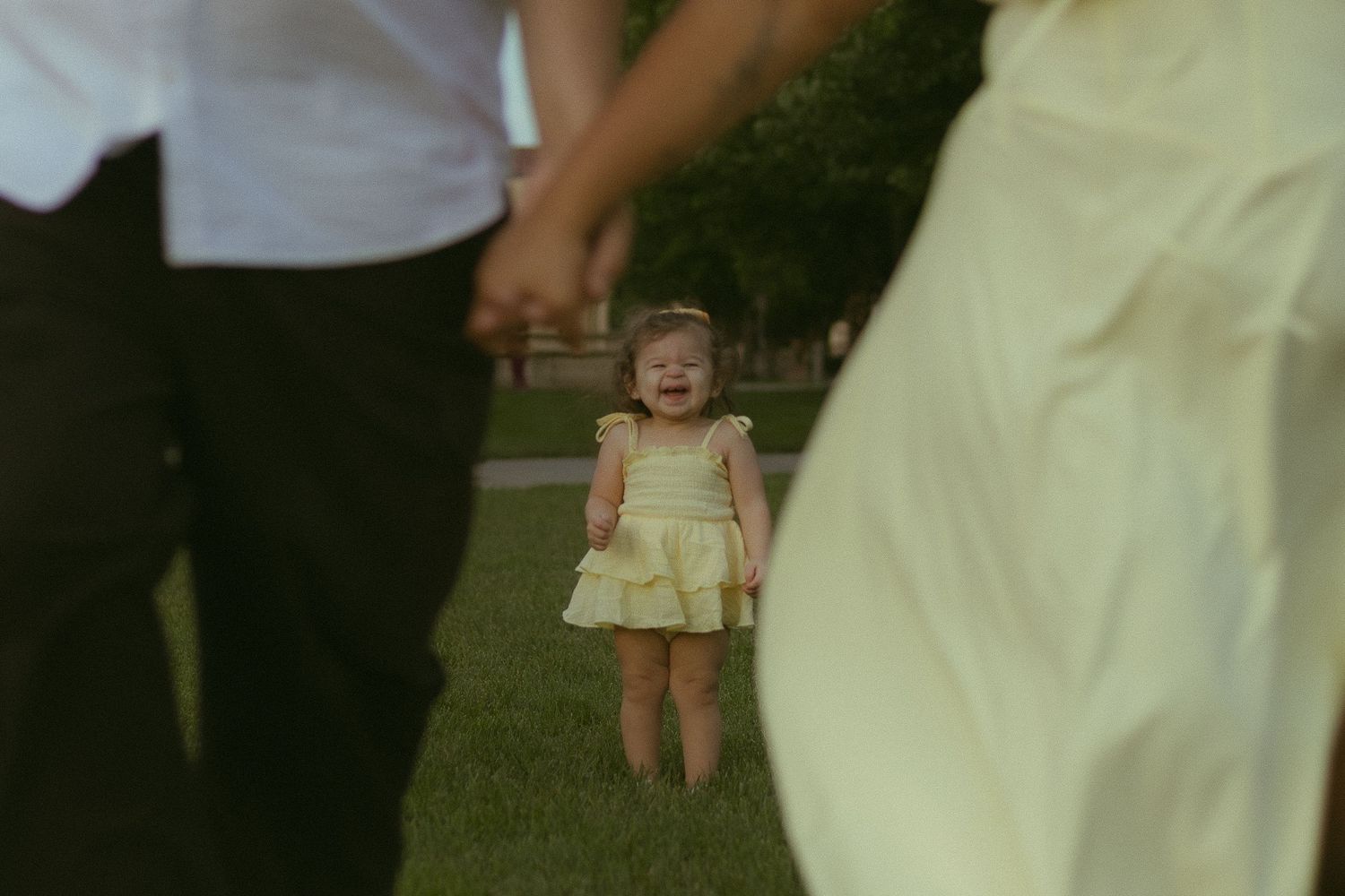 A toddler girl in a yellow dress holds her parents hands while standing on green grass outdoors.
