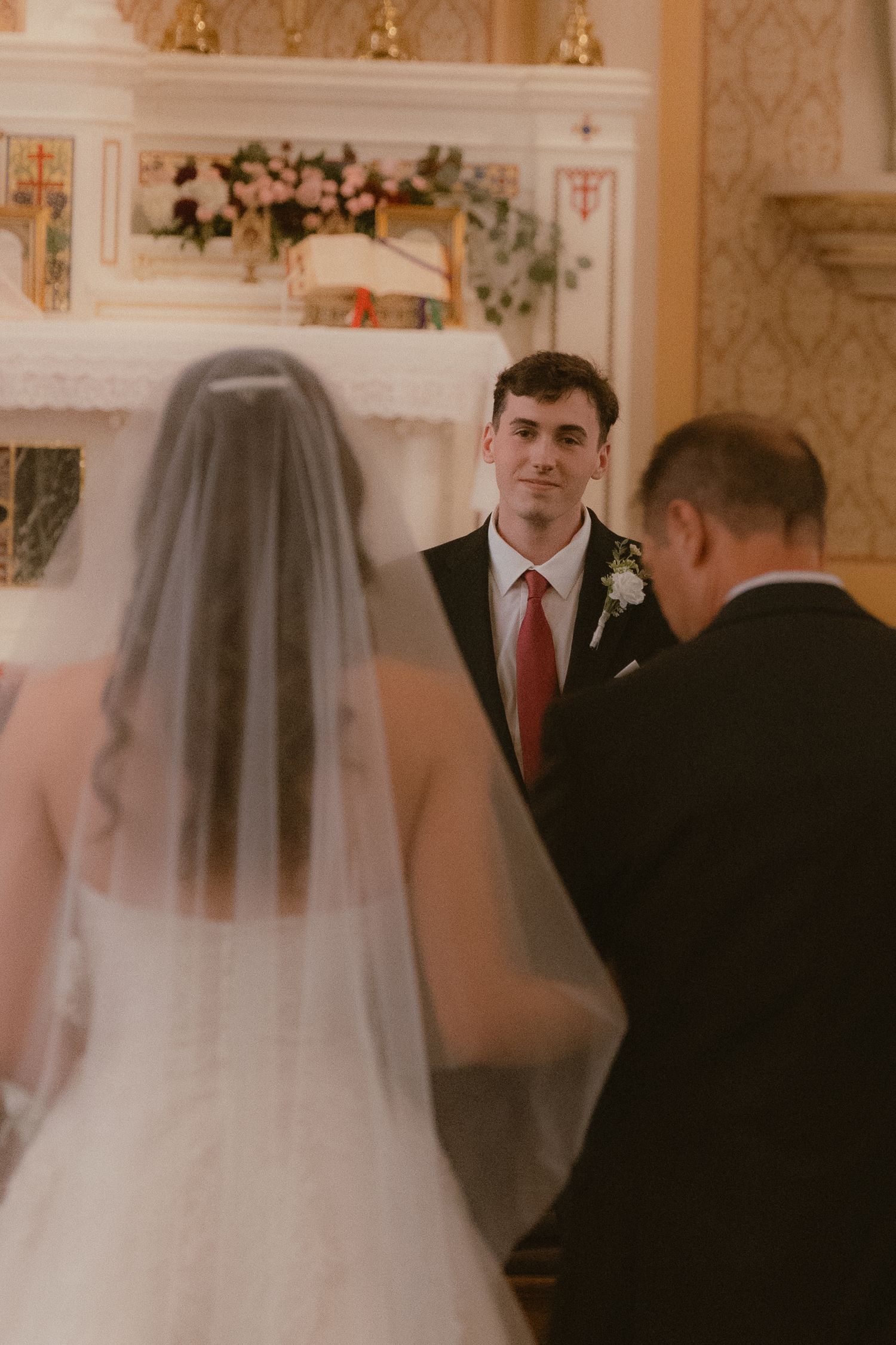 Groom in black suit with red tie smiling at bride during church wedding ceremony at decorated altar.