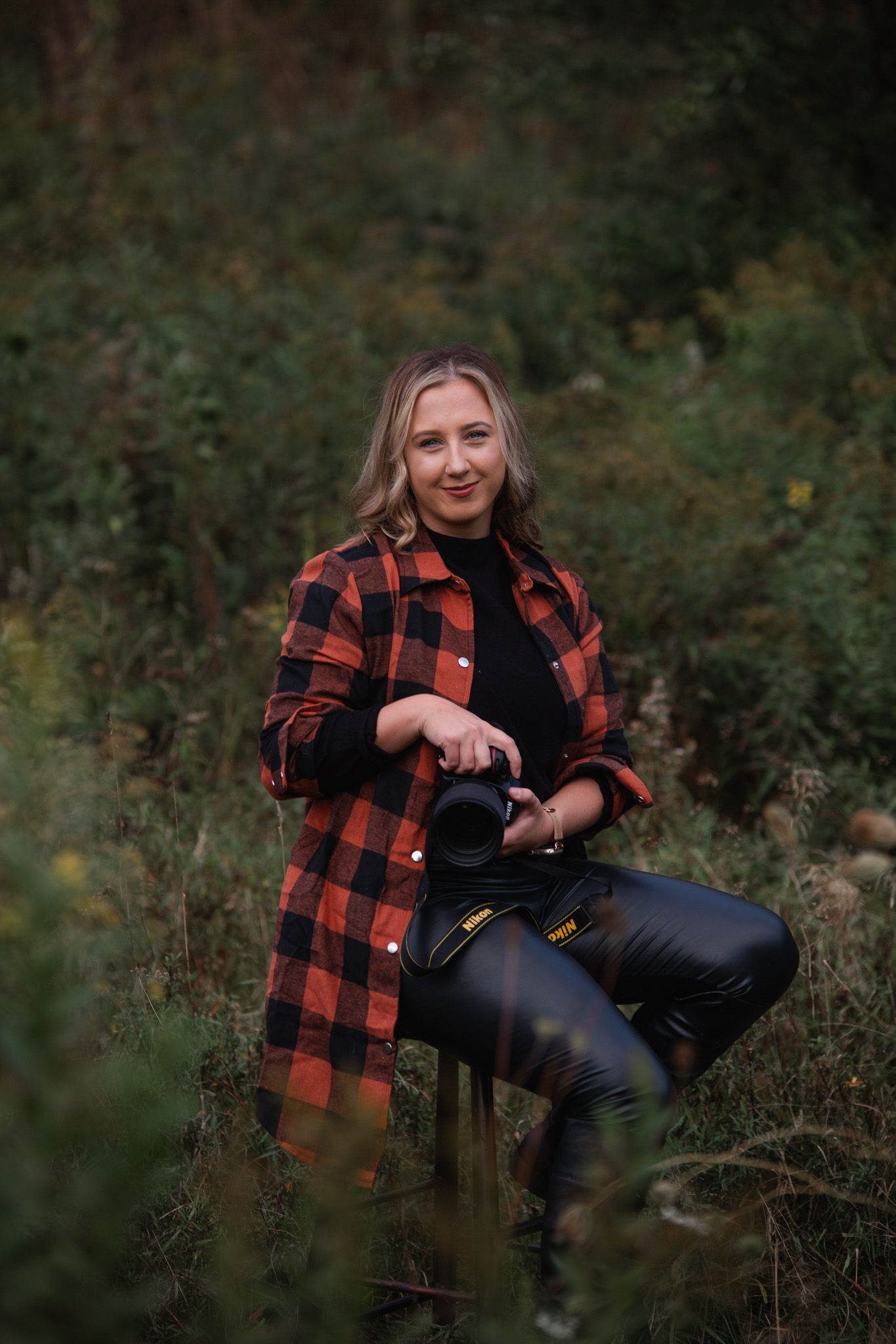 Person wearing red buffalo plaid flannel and black pants poses with camera in outdoor autumn setting.
