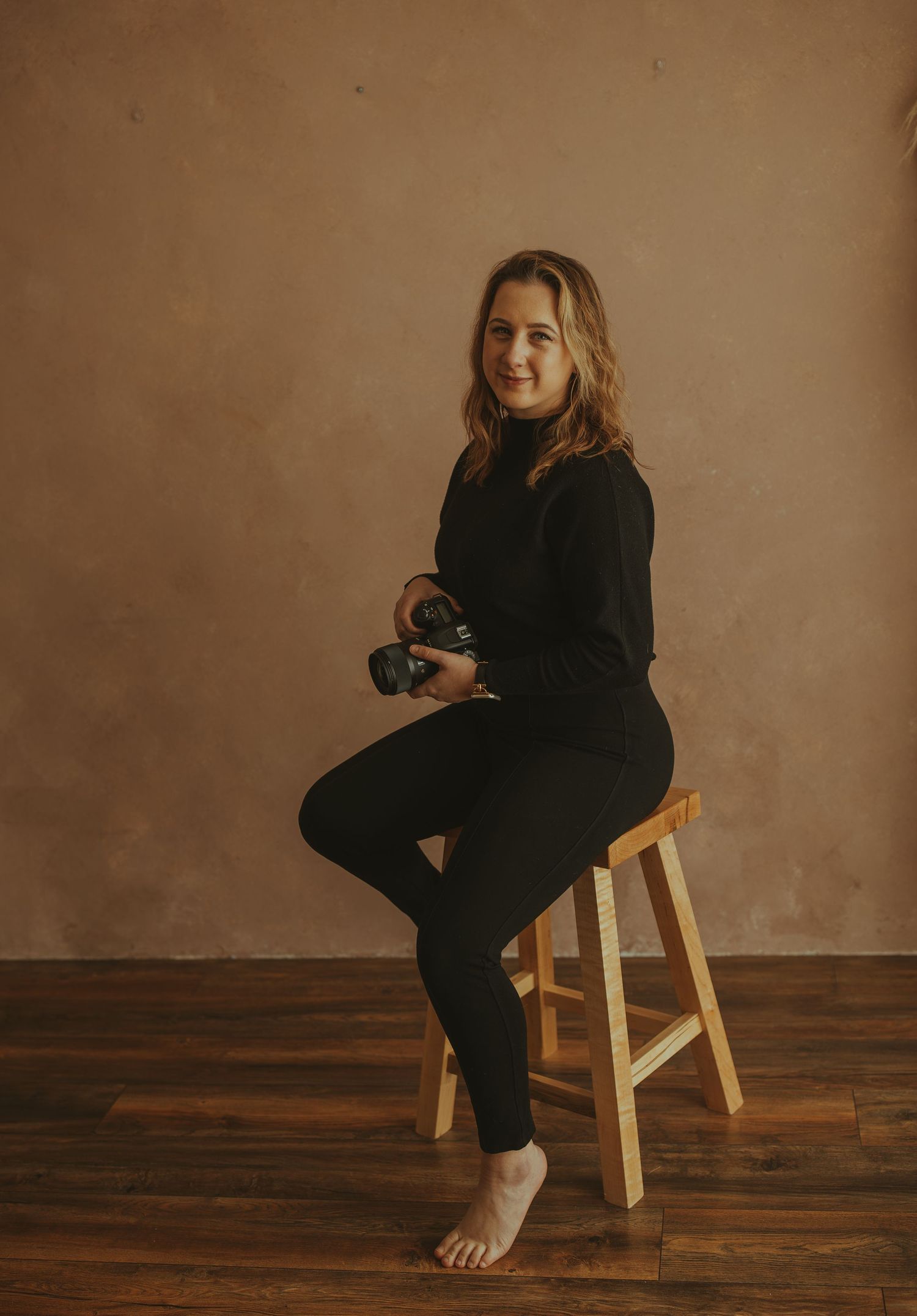 Professional photographer sits on wooden stool wearing all black while holding camera against brown backdrop.