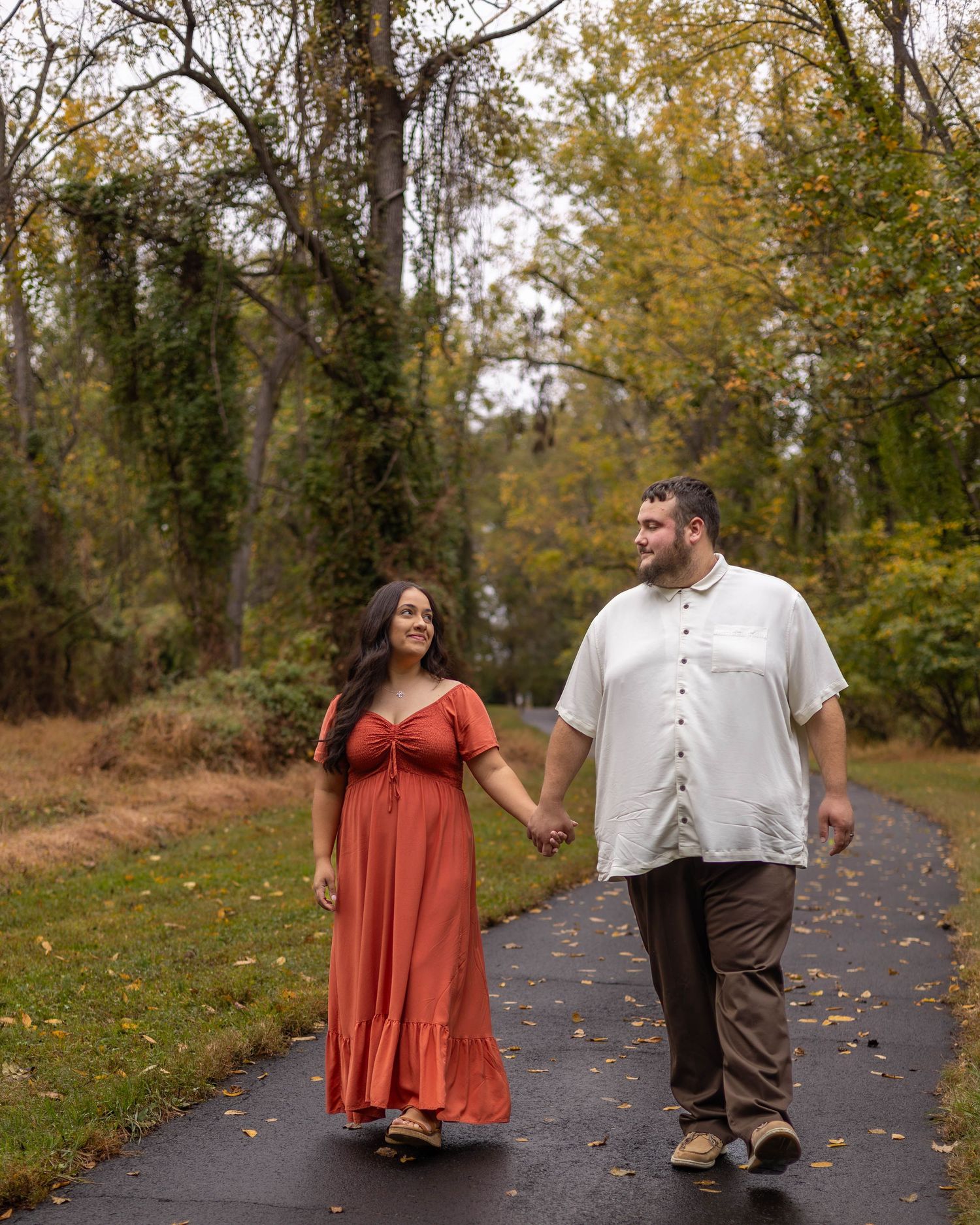 Two people walk together on a paved trail surrounded by autumn trees with fallen leaves on the ground.