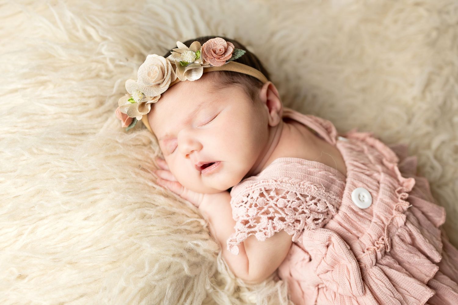 Sleeping newborn in pink dress and headband lying on cream-colored fur blanket.