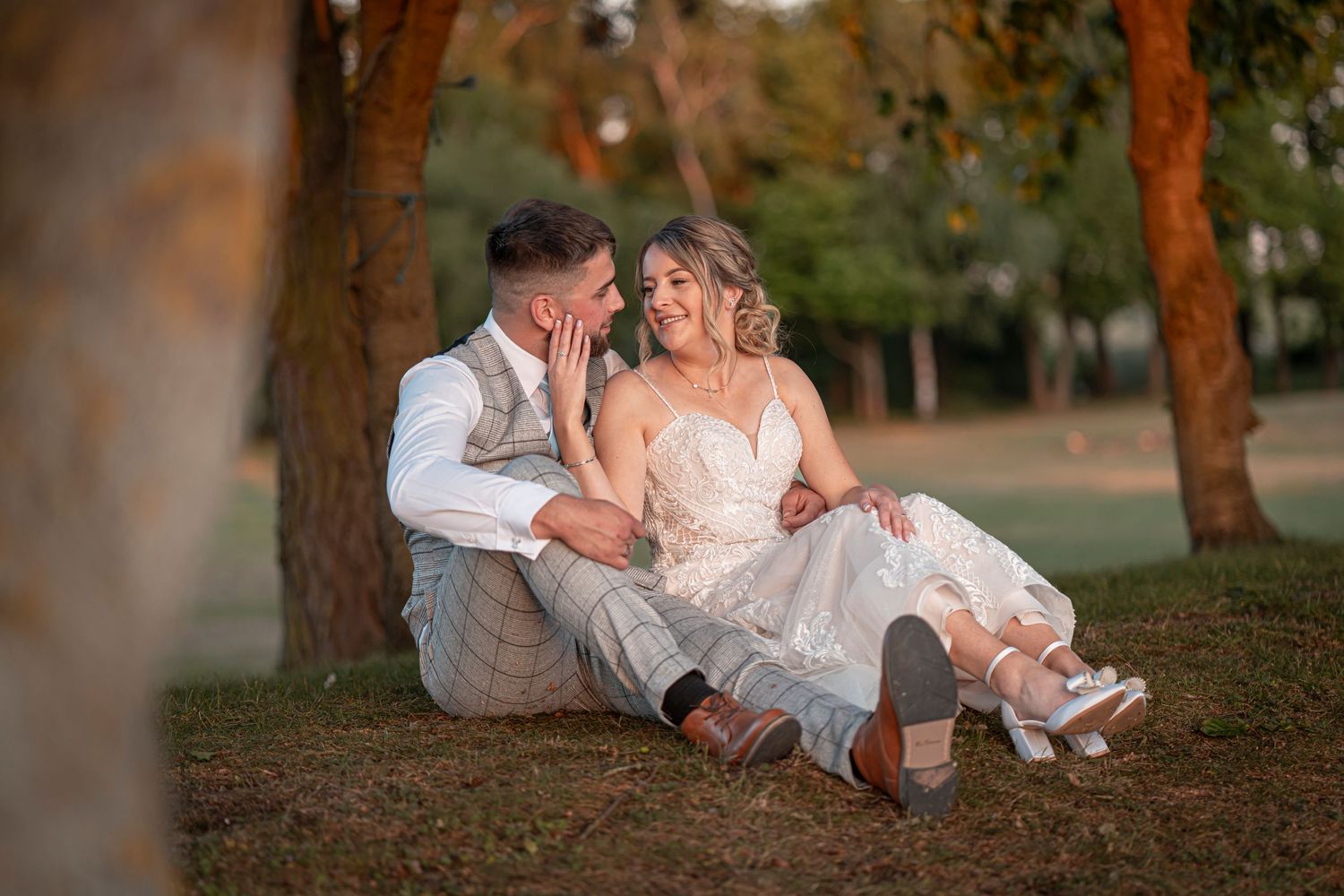 Casual wedding portrait of couple relaxing under trees in warm lighting.