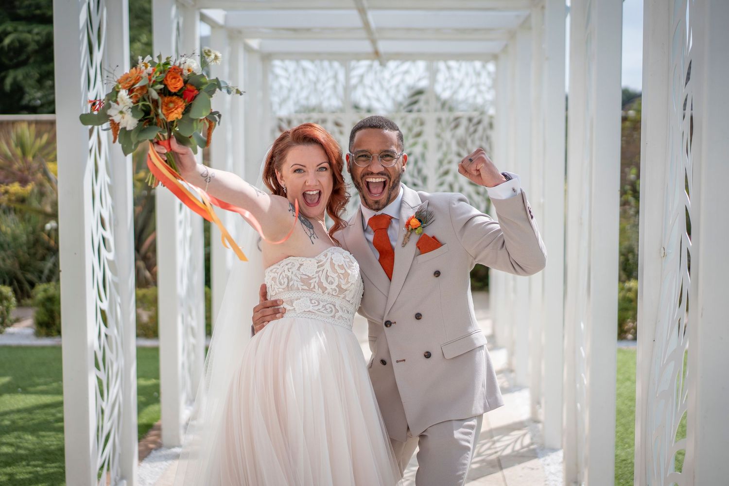 Newlyweds share a joyful moment under a white pergola decorated with orange floral accents.