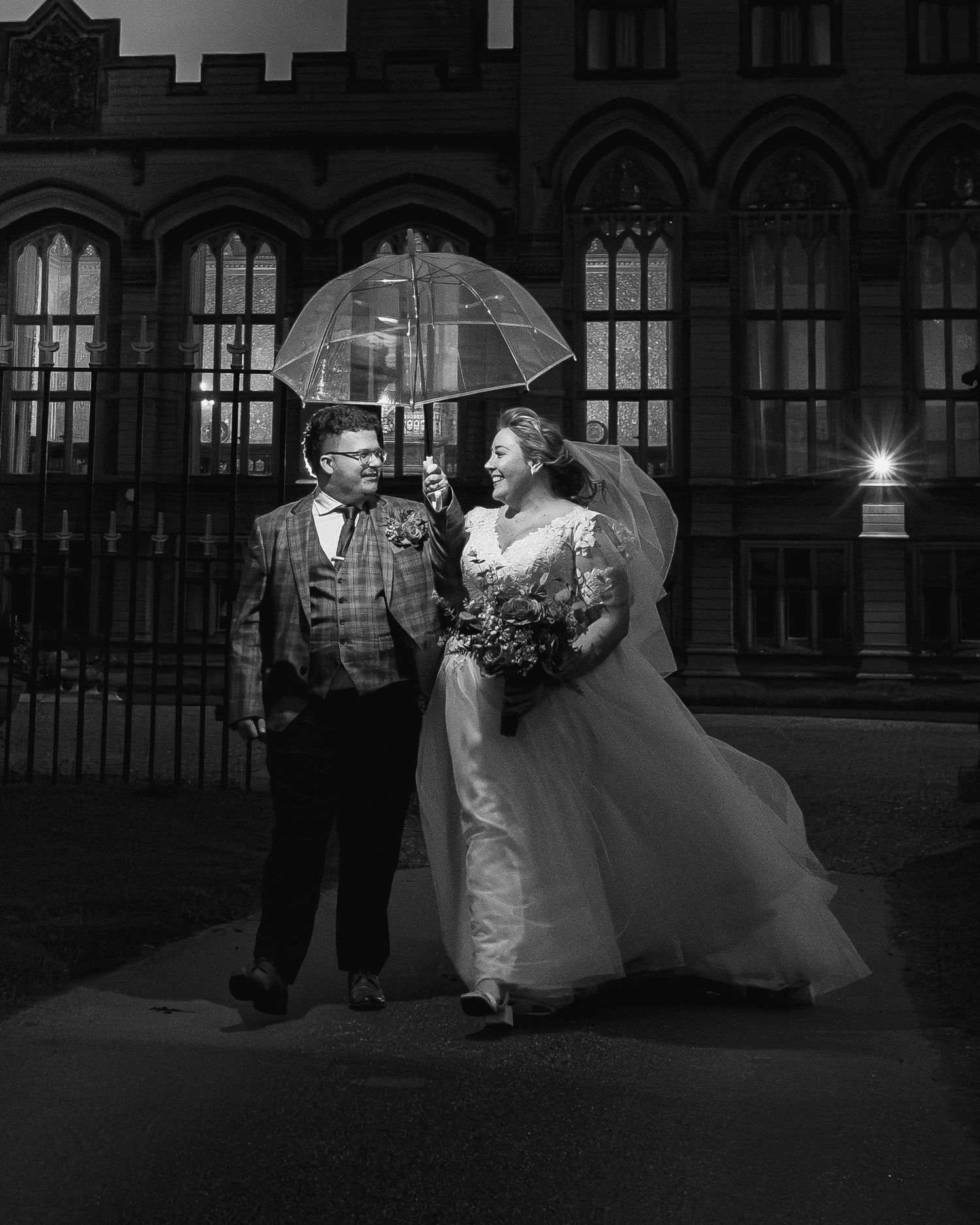 A romantic black and white wedding photo with a couple sharing an umbrella against Gothic architecture.