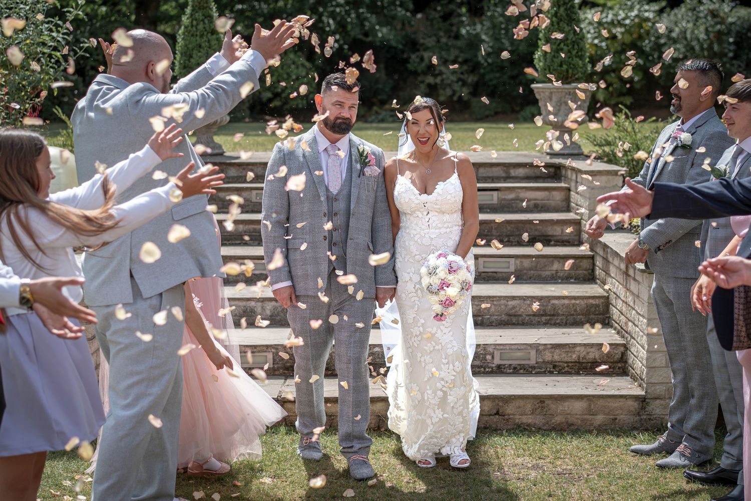 Wedding party walking down outdoor steps while guests throw flower petals in celebration.