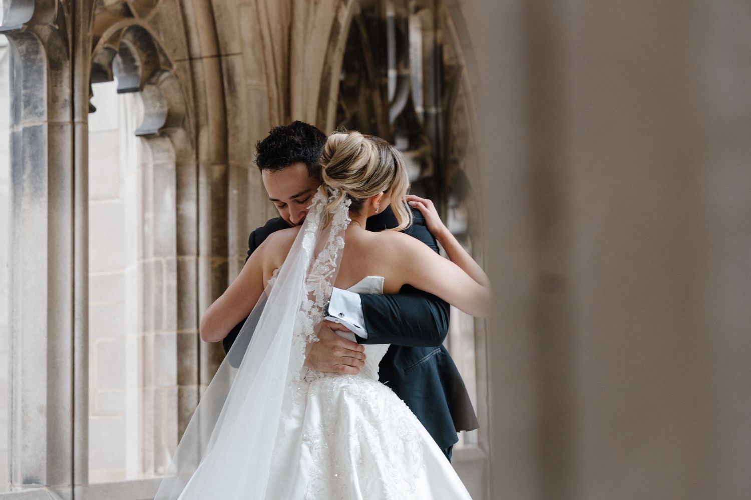Bride and groom share a tender embrace in an elegant stone archway, bride wearing a lace gown and veil.