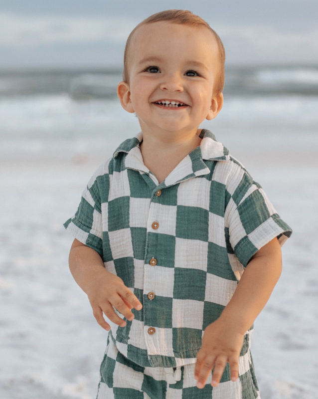 Toddler wearing green and white checkered shirt smiles joyfully at the beach.
