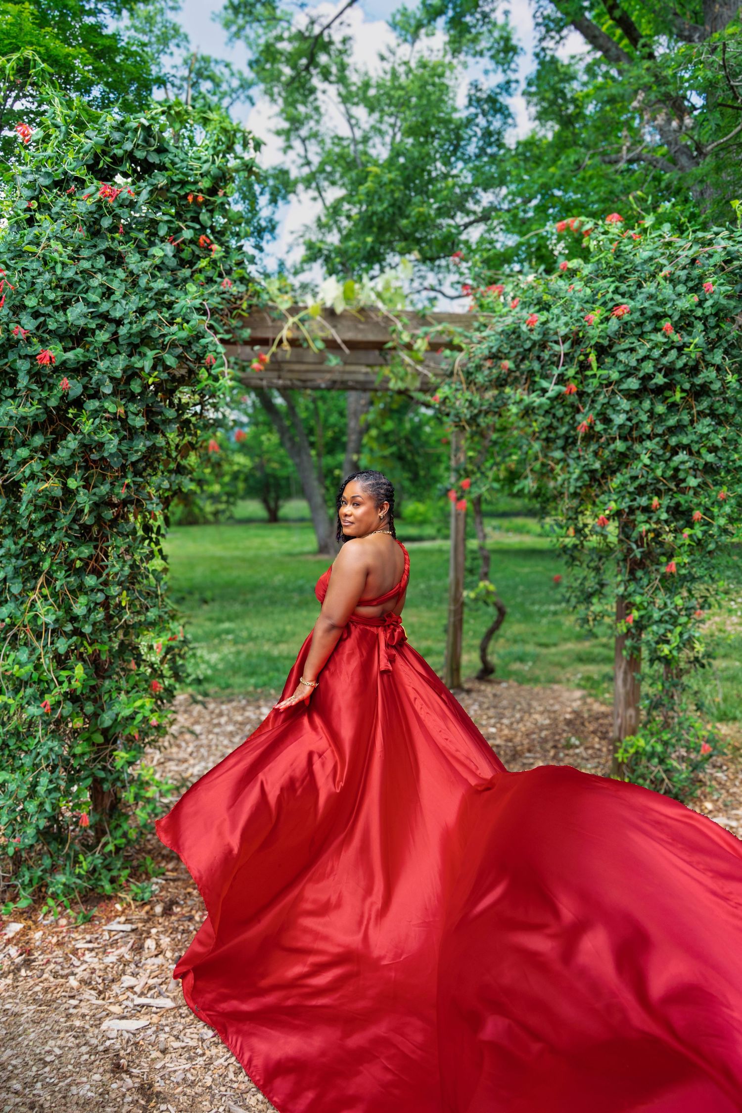 Woman in flowing red gown poses gracefully under a vine-covered garden arbor with lush greenery.