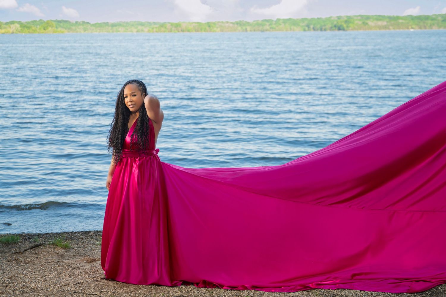 Long flowing fuchsia dress billows in the wind against a serene lakeside backdrop with blue water and lush greenery.