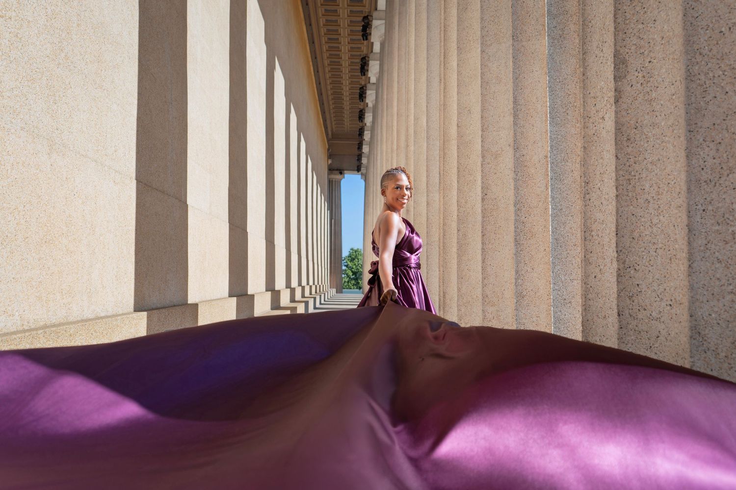 Woman in flowing purple gown poses among tall stone columns in an elegant architectural setting.