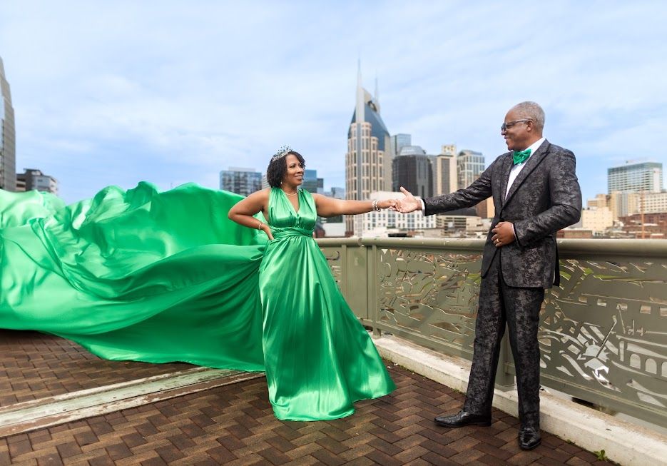 Couple in matching green outfits poses on Nashville rooftop, woman's flowing gown billowing in wind with city skyline behind them.
