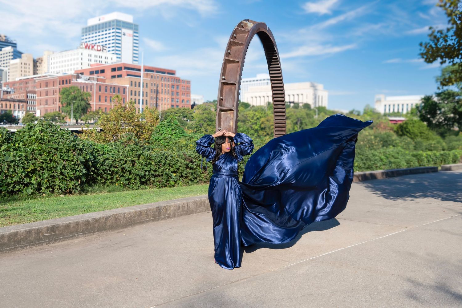 Woman in flowing blue satin gown poses dramatically before large circular sculpture with city skyline backdrop.