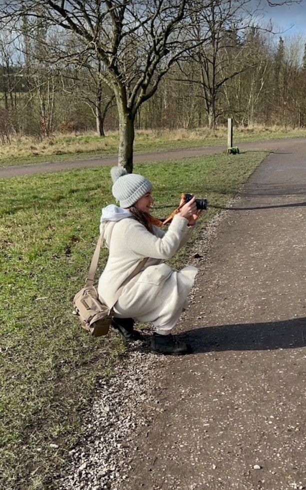 Woman in a cream hoodie and gray knit beanie crouches on a gravel path, taking a photo with a camera beside a grassy field and bare trees.