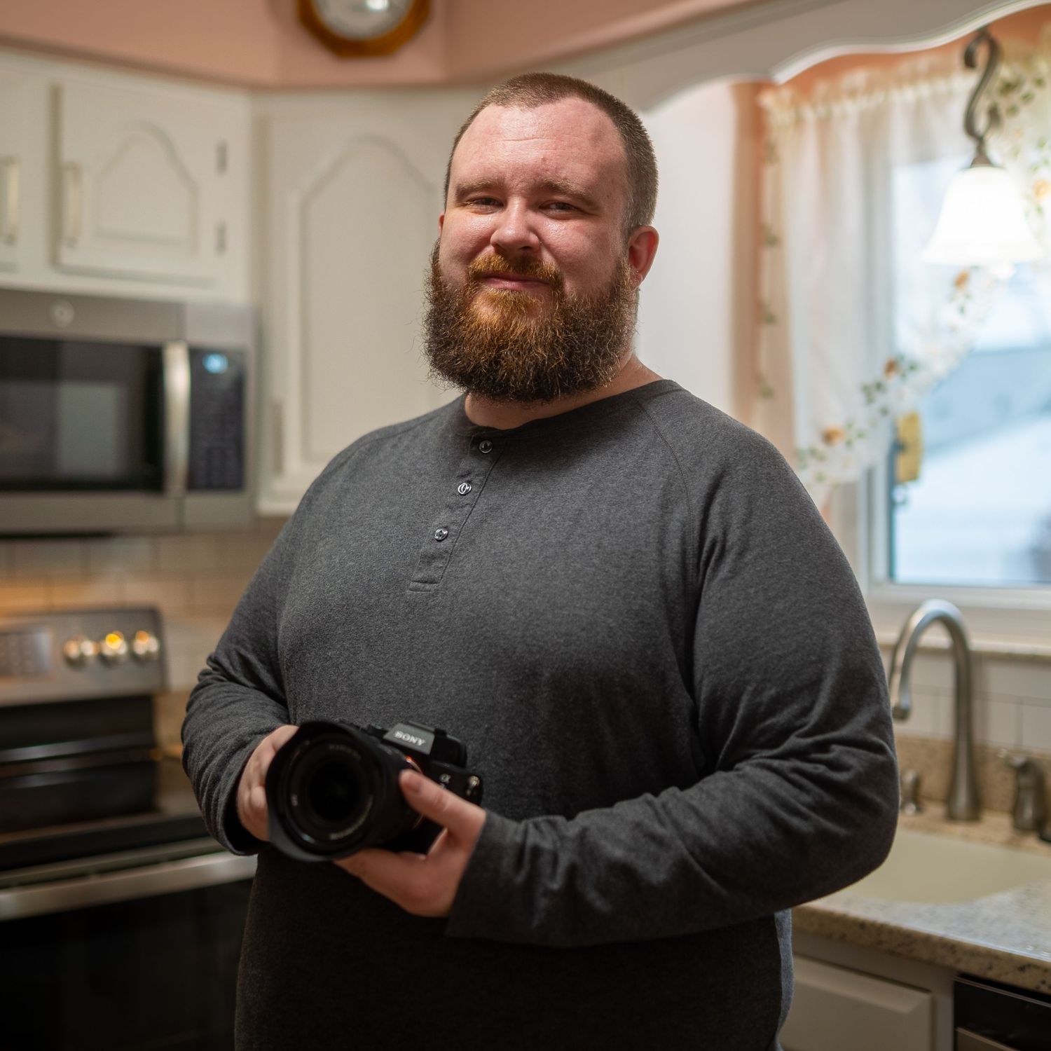 Bearded man holding a camera smiling in a modern kitchen with white cabinets and stainless steel appliances.
